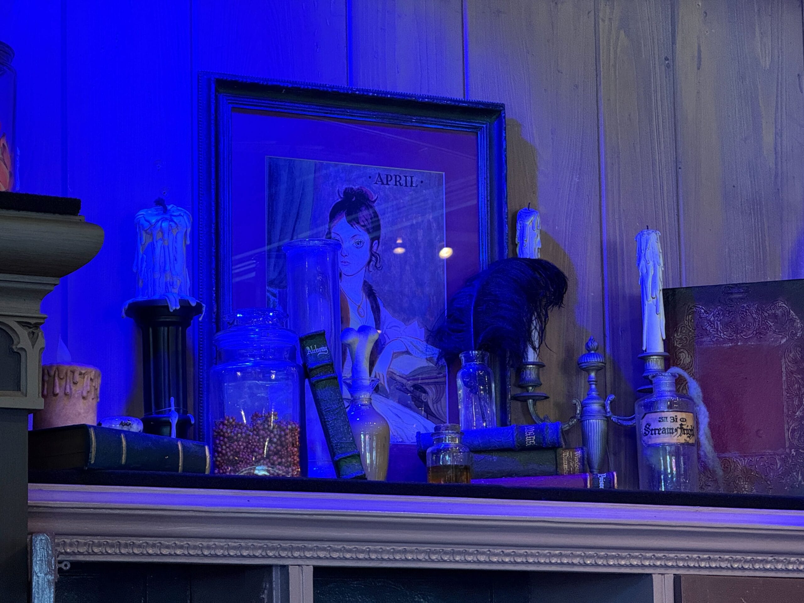 Shelf with old books, jars, candlesticks, and a framed "April"—like a Haunted Mansion Store display—under blue lighting.