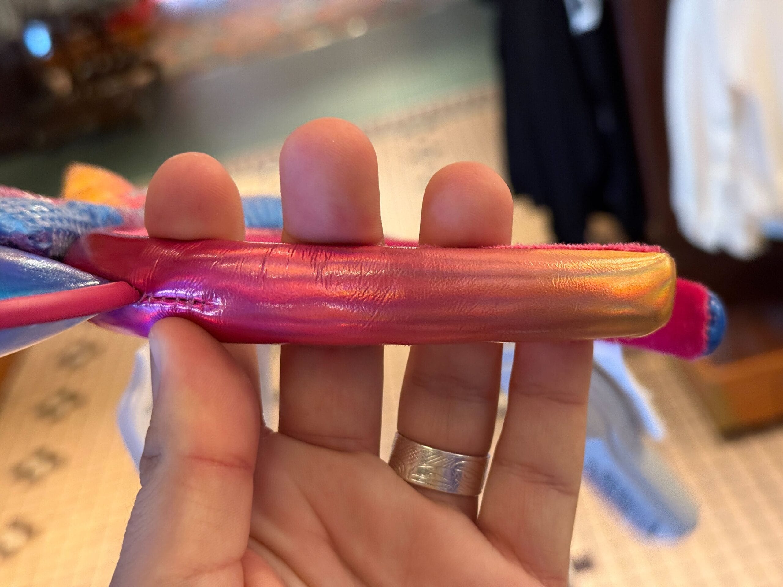 A hand holds a shiny, iridescent pink and orange strap, with blurred clothing in the background.