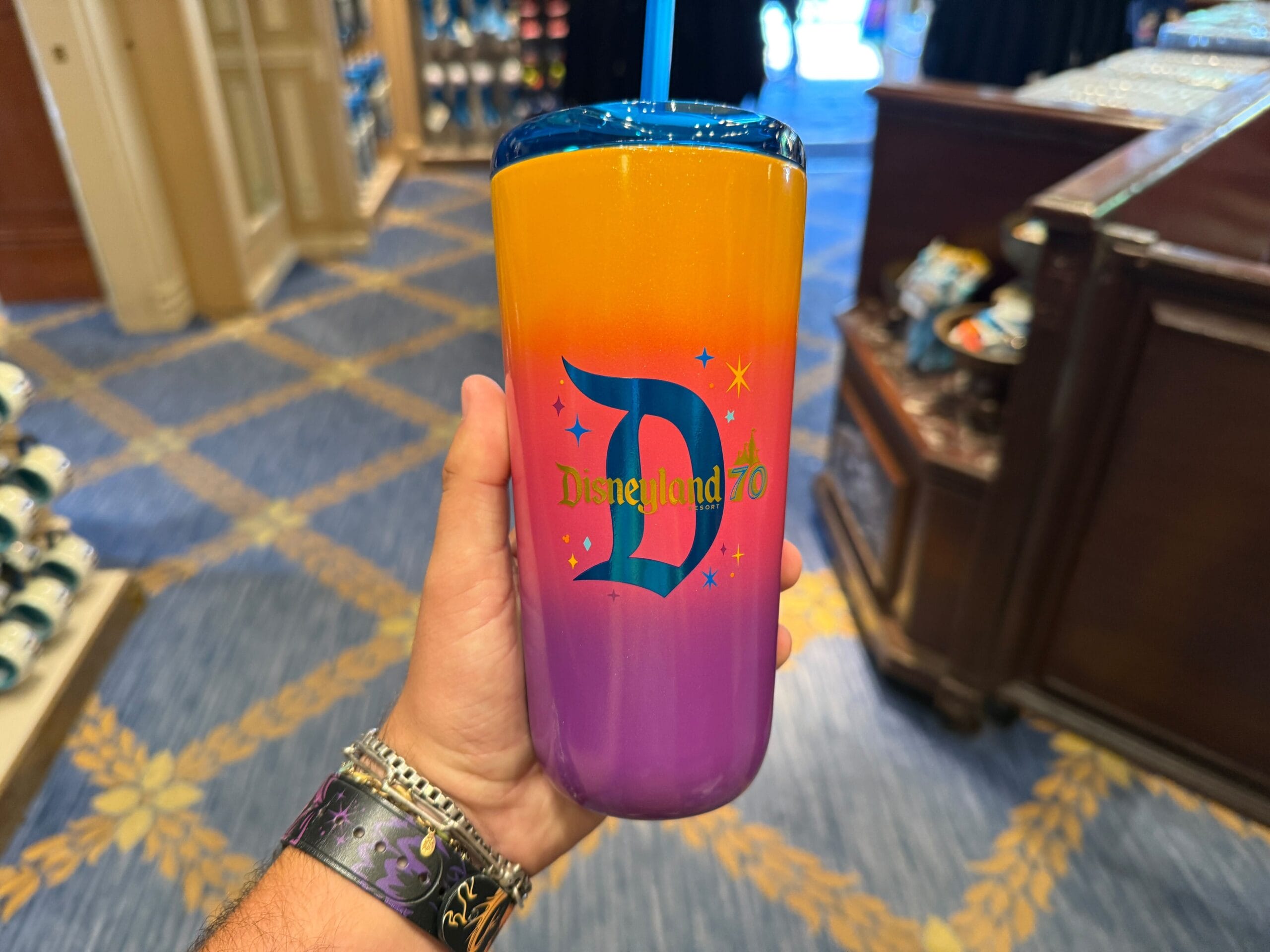 A hand holds a colorful Disneyland 70th Anniversary tumbler beside a Loungefly Backpack in a store with blue and gold carpet.