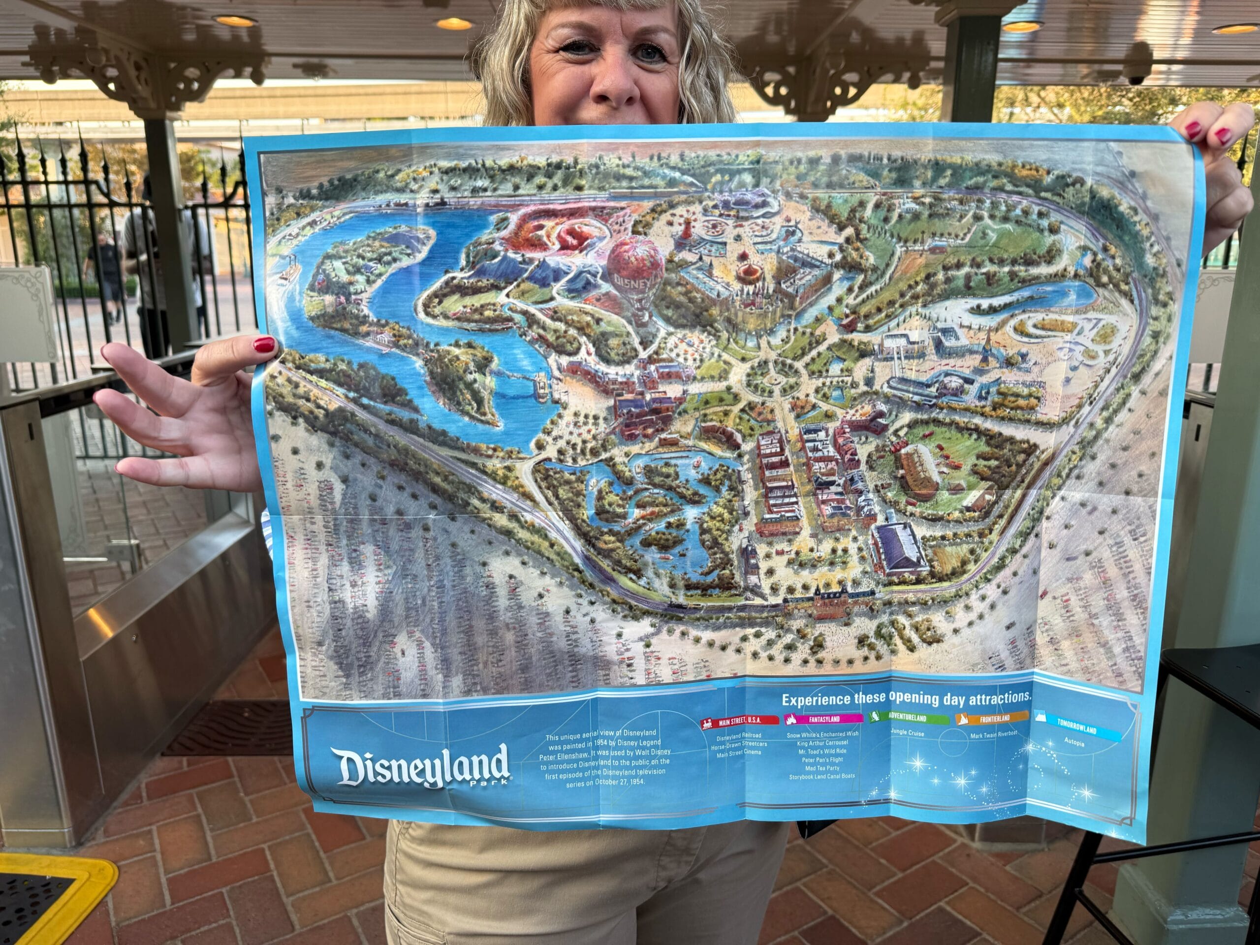 A woman holds up a detailed illustrated map of Disneyland at an entrance gate, showing colorful park attractions.