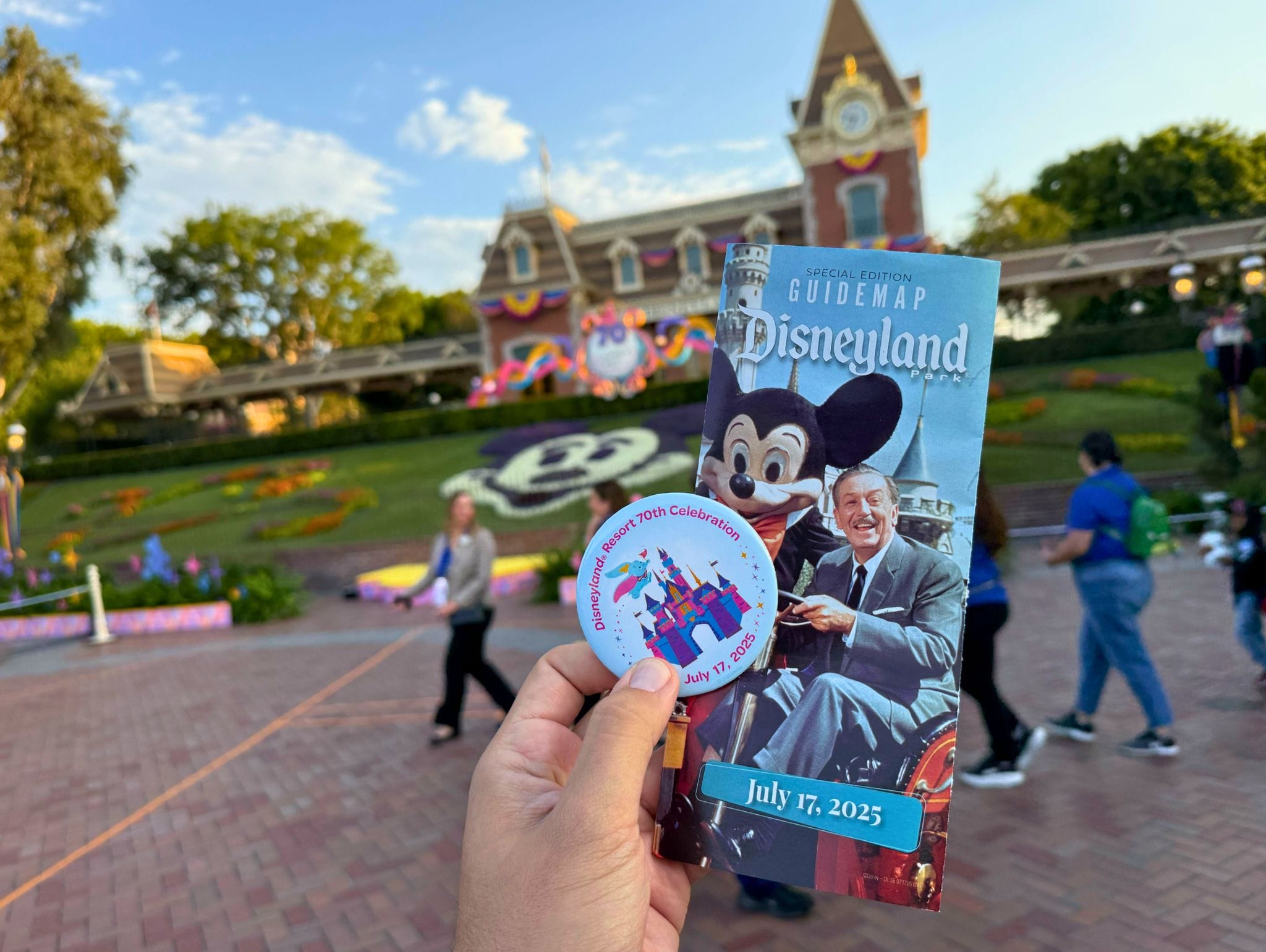 Hand holding a Disneyland guidemap and a 70th celebration button in front of Disneyland’s entrance.