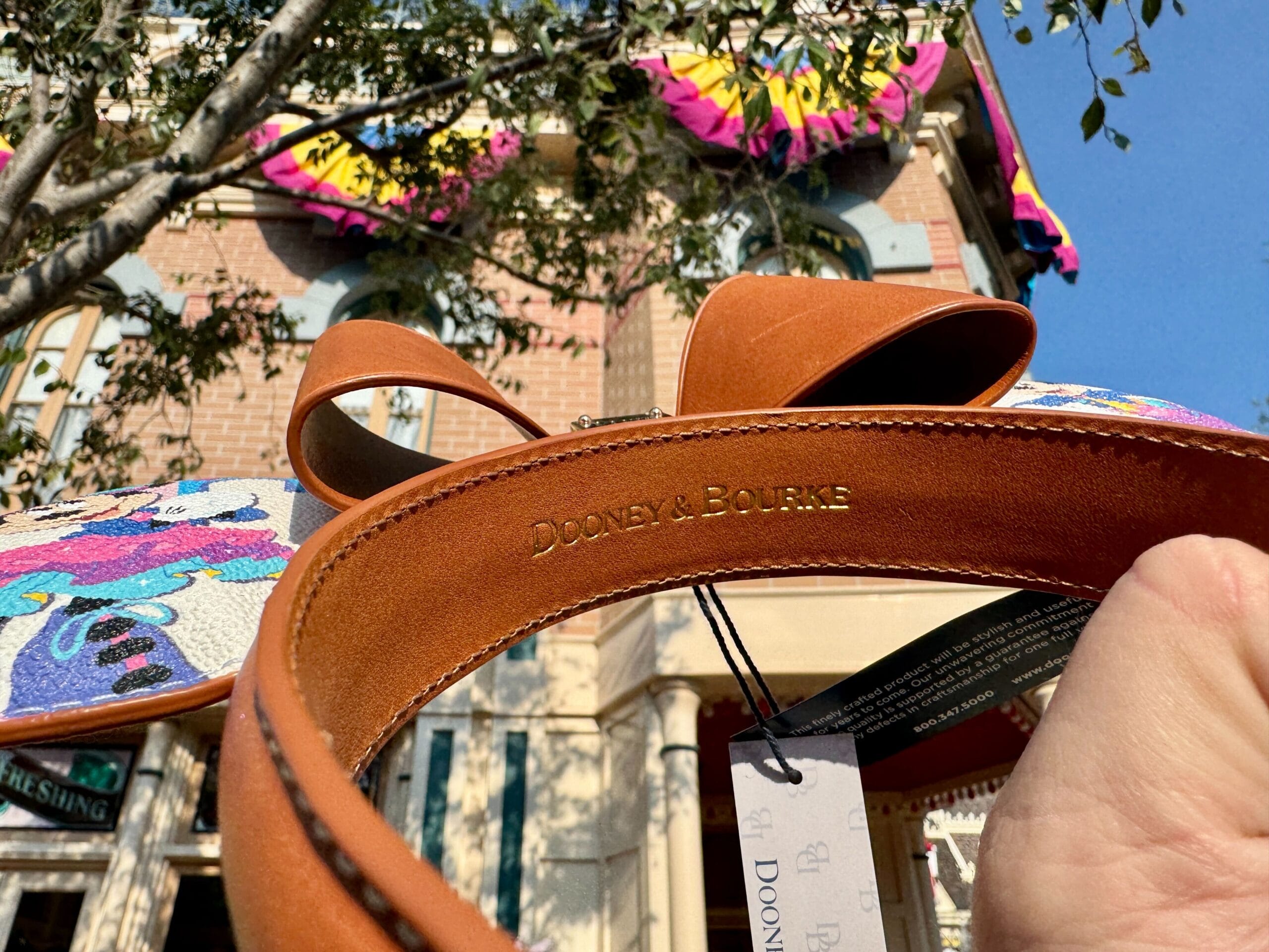A hand holds Disneyland 70th Anniversary merchandise—Mickey Mouse ears—outside a brick building with colorful banners.