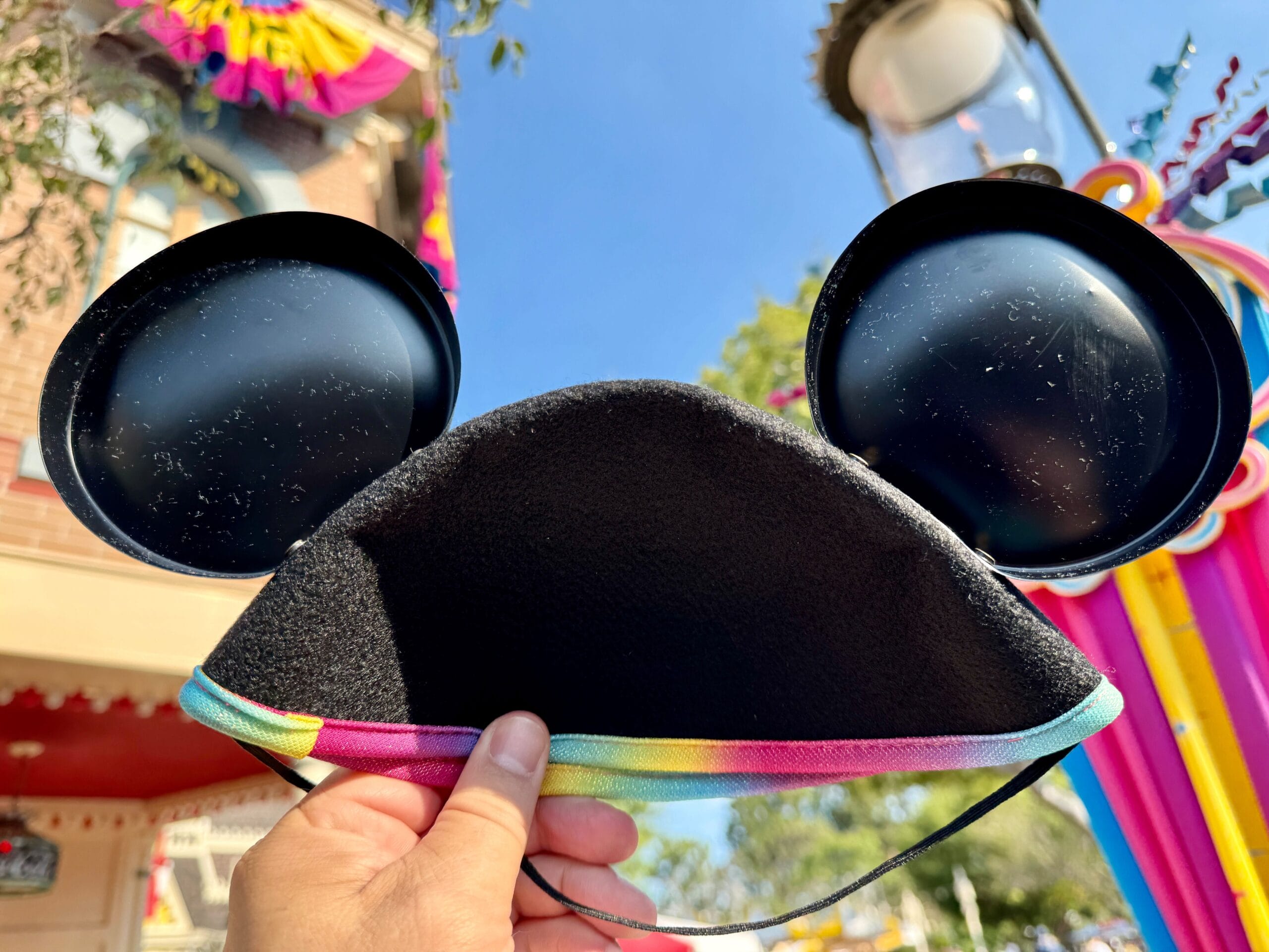 A hand holds up Disneyland 70th Anniversary Mickey Mouse ears hat with rainbow trim outside under a clear blue sky.