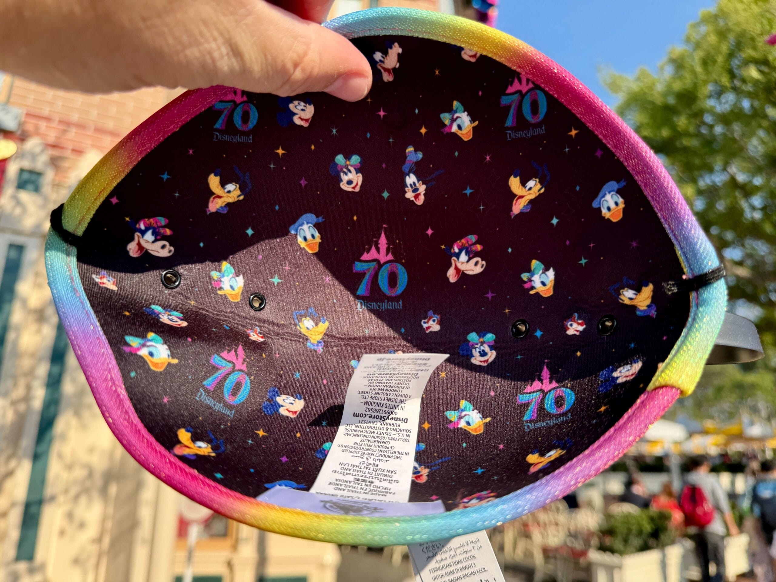 Hand holding Disneyland 70th Anniversary merchandise hat with Disney characters and rainbow edge, outdoors.