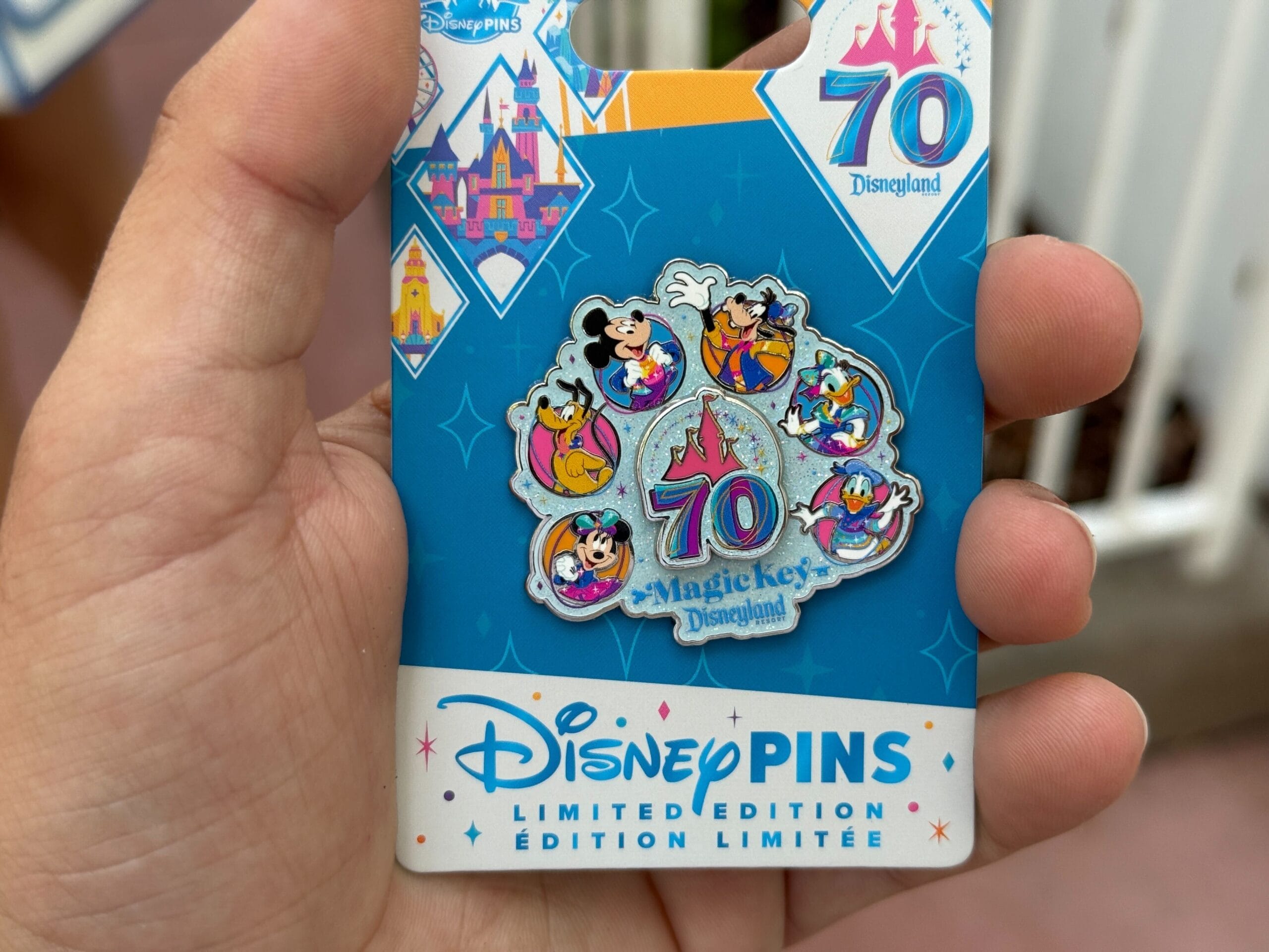 A hand holds a Disneyland 70th Anniversary Magic Key pin, showcasing classic characters on blue merchandise packaging.