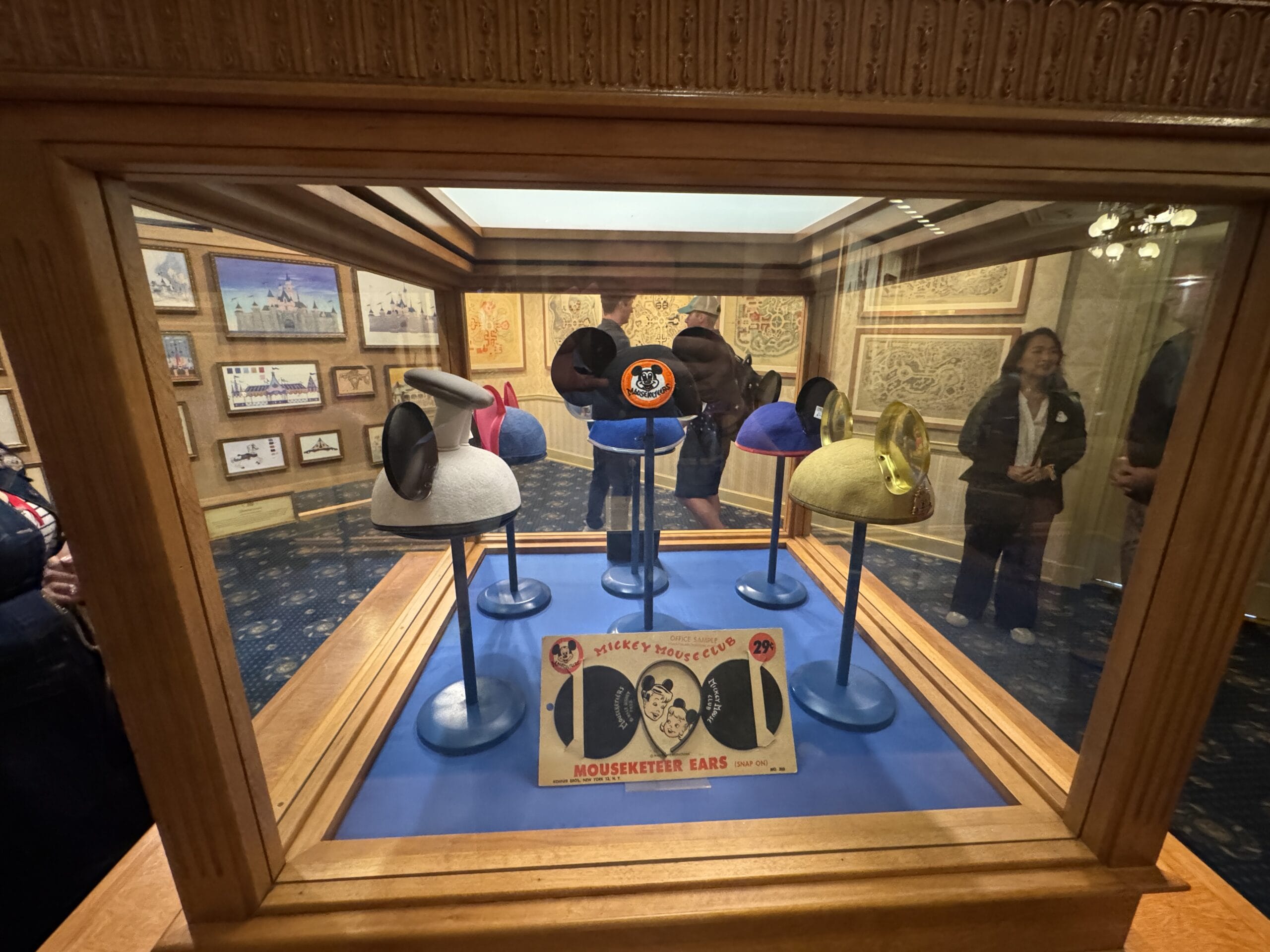 A Main Street Opera House display case features vintage Mickey Mouse ear hats and a Mouseketeer sign, with visitors viewing photos.
