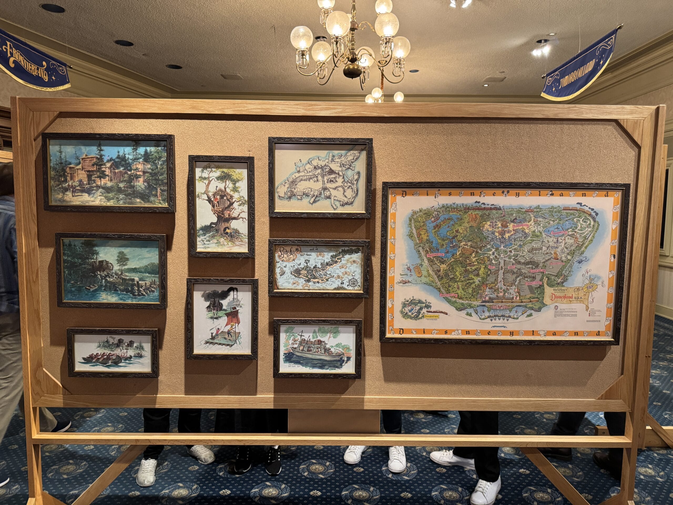 A display board with framed vintage Disneyland maps, artwork, and photos; legs of people at the Main Street Opera House visible.