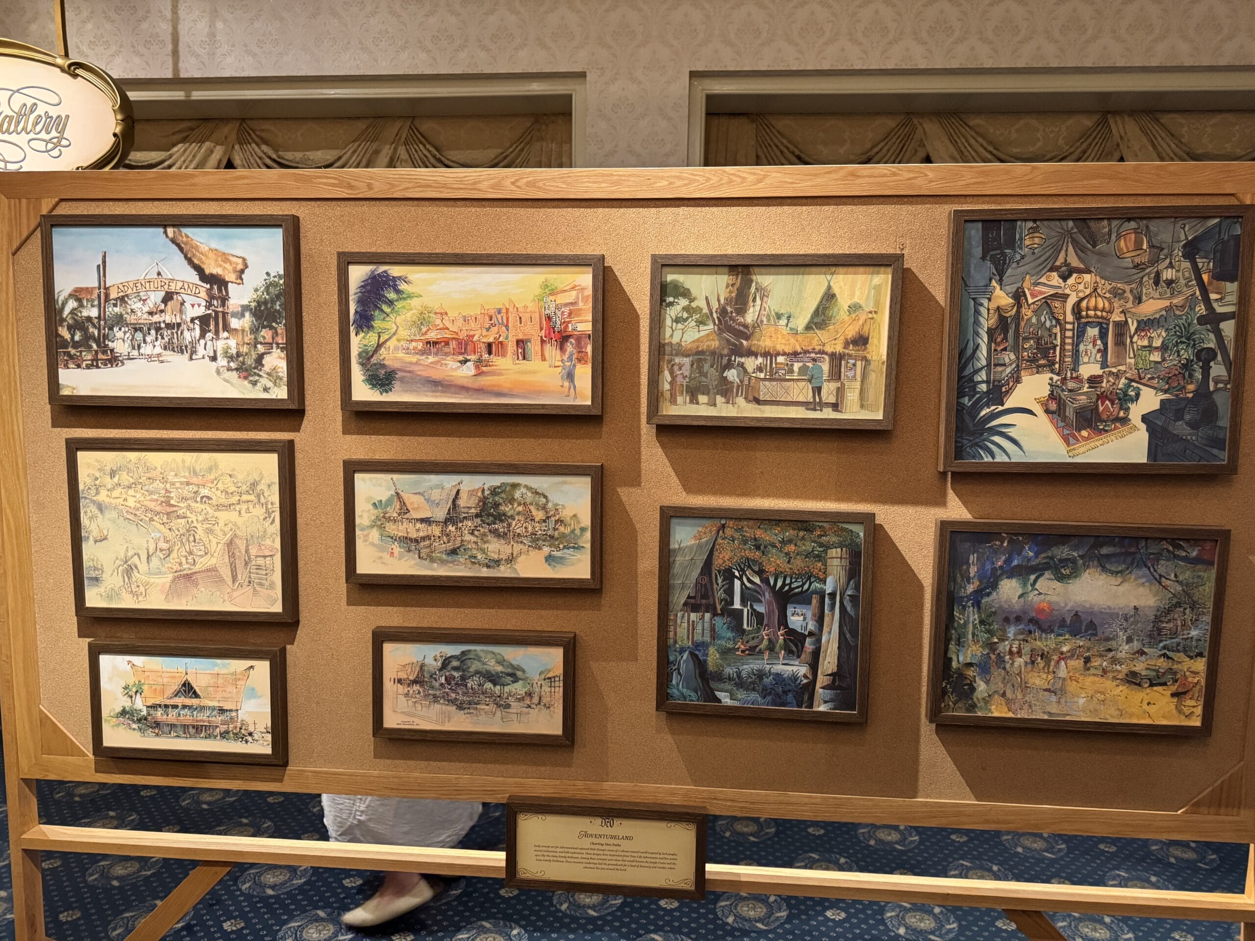 A display board at Main Street Opera House holds nine framed paintings depicting vibrant, adventure-themed village scenes.