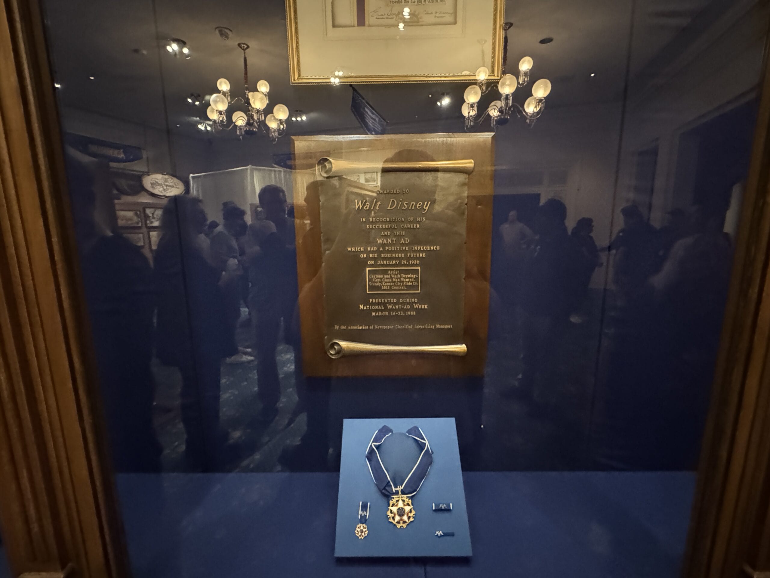 A medal and plaque honoring Walt Disney are displayed at the Main Street Opera House, with people reflected in the glass.