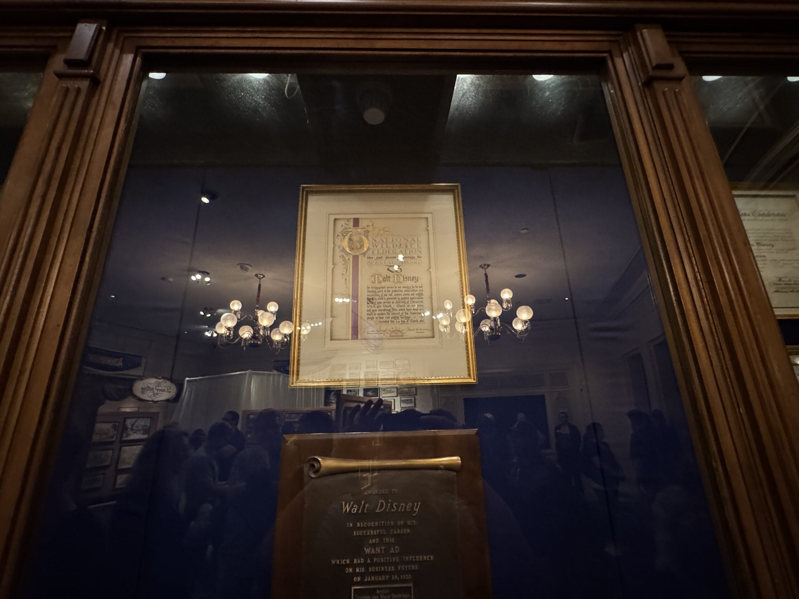 Framed certificate and plaque for Walt Disney at Main Street Opera House, reflected crowd visible in the glass.
