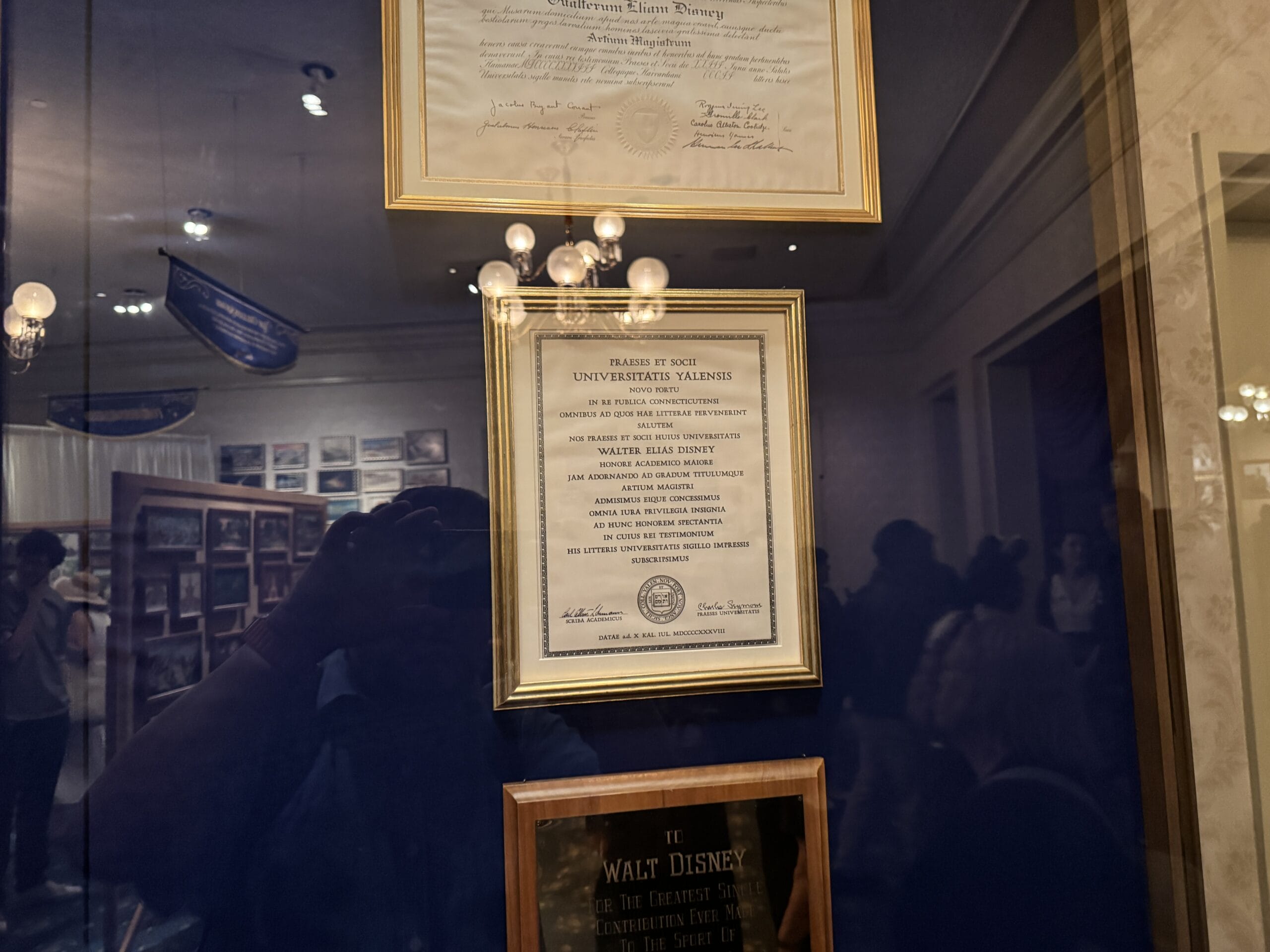 Framed diploma displayed on a wall at Main Street Opera House, with reflected lights and people in the glass.