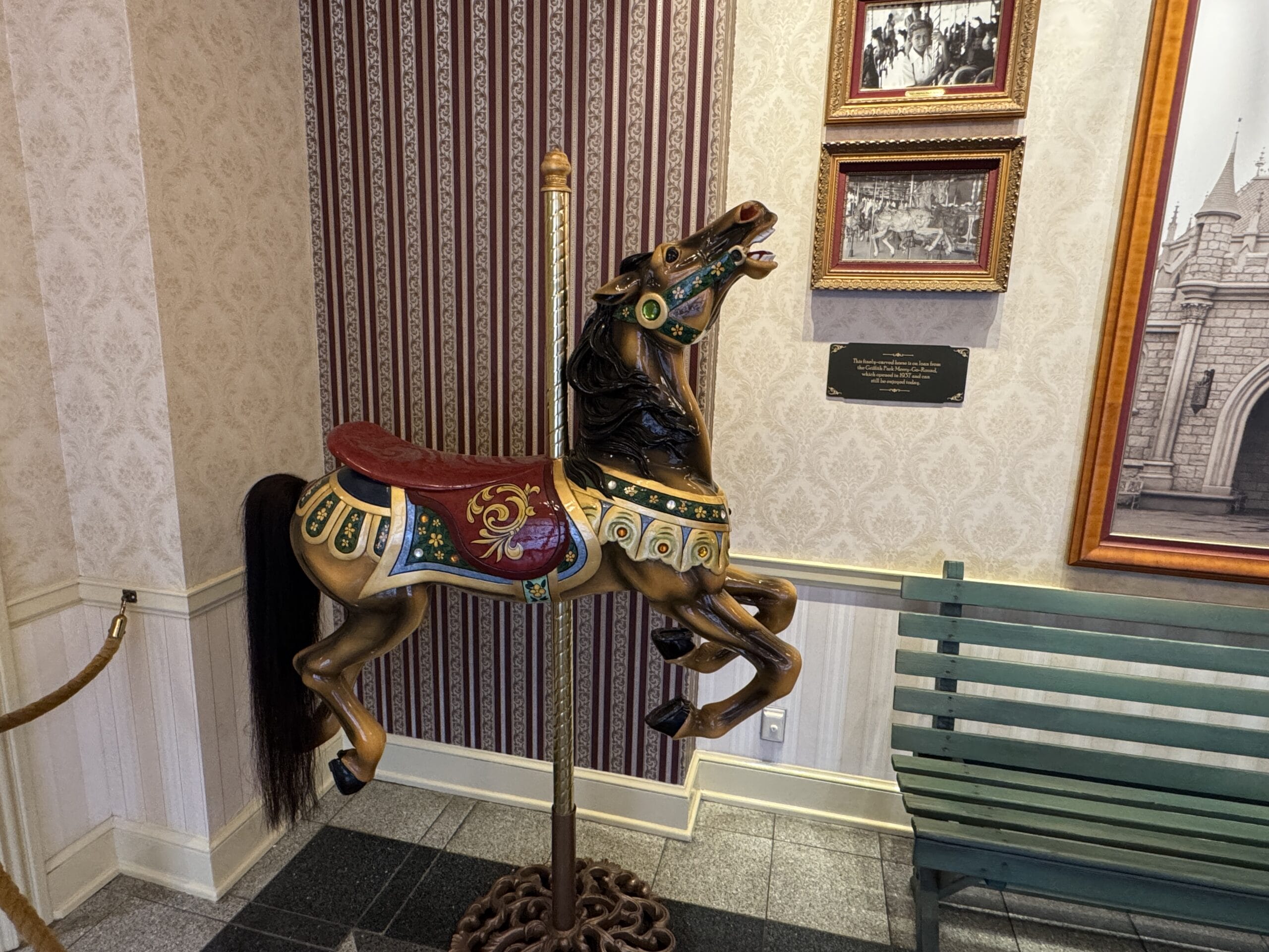 A colorful carousel horse, echoing Disneyland history, sits indoors beside a green bench and framed photos on a patterned wall.