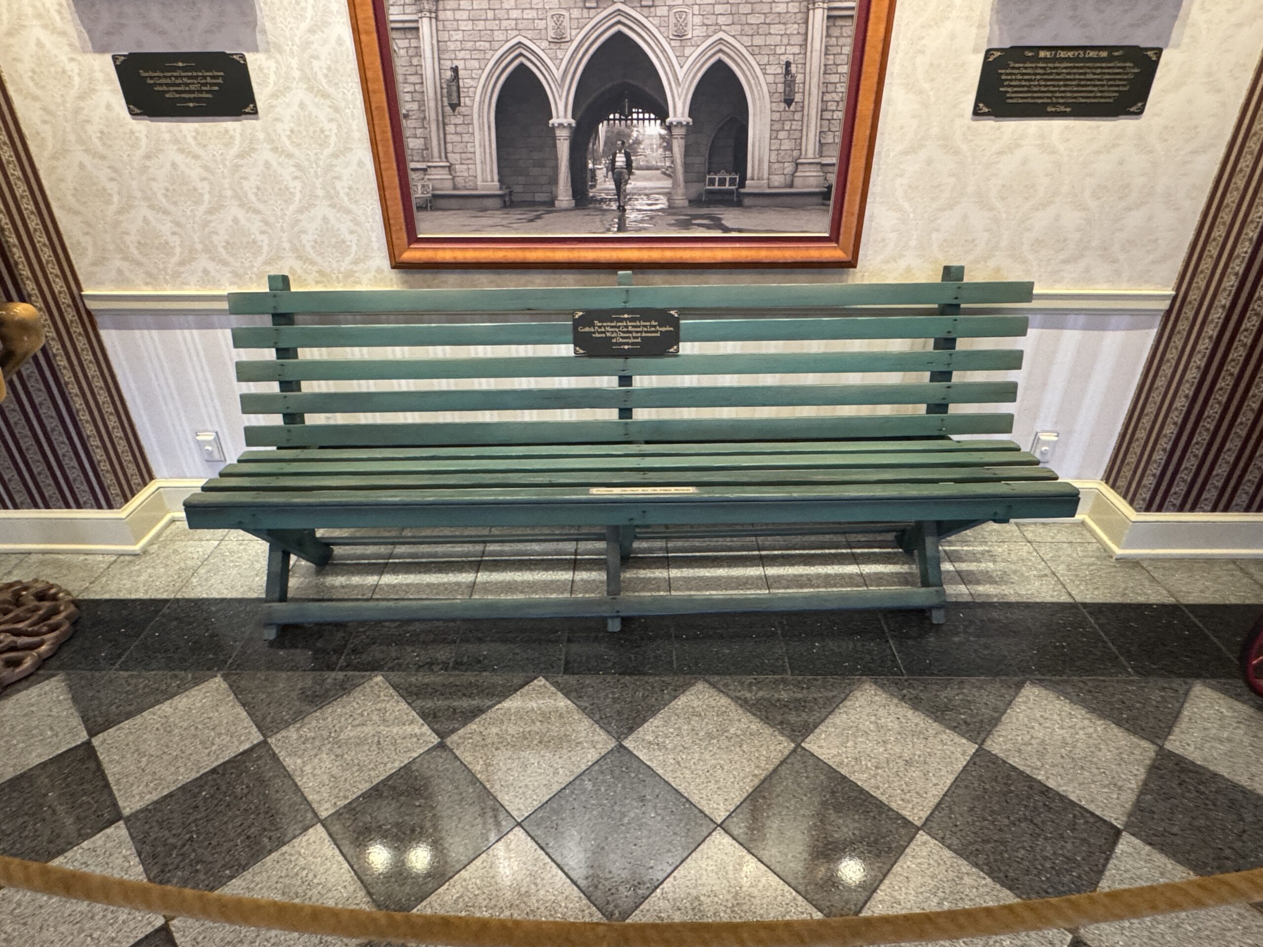 A green wooden bench from Main Street Opera House, displayed indoors with plaques, framed photo, and patterned wallpaper.