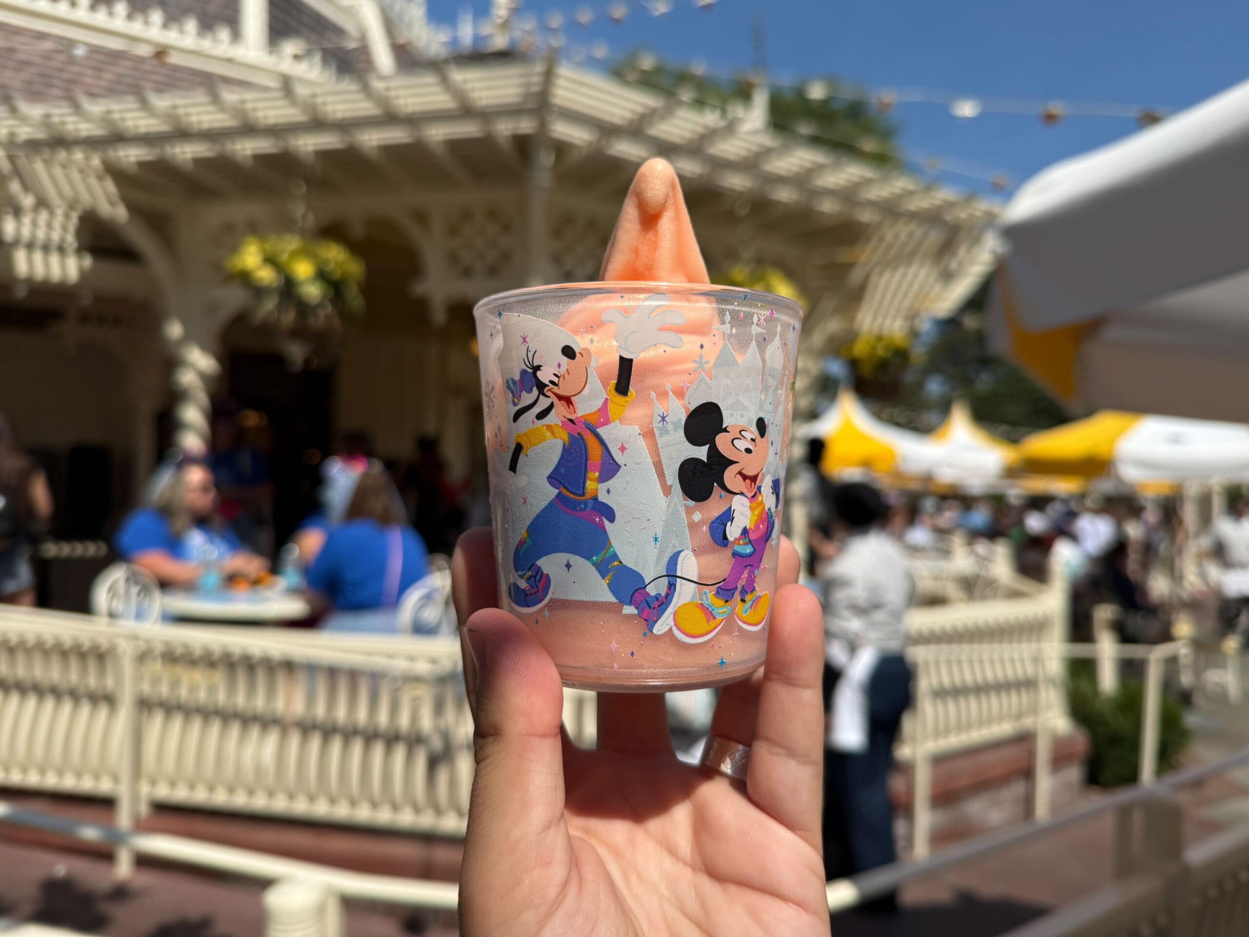A hand holds a Disney-themed cup with a pink sherbet treat in front of an outdoor café.