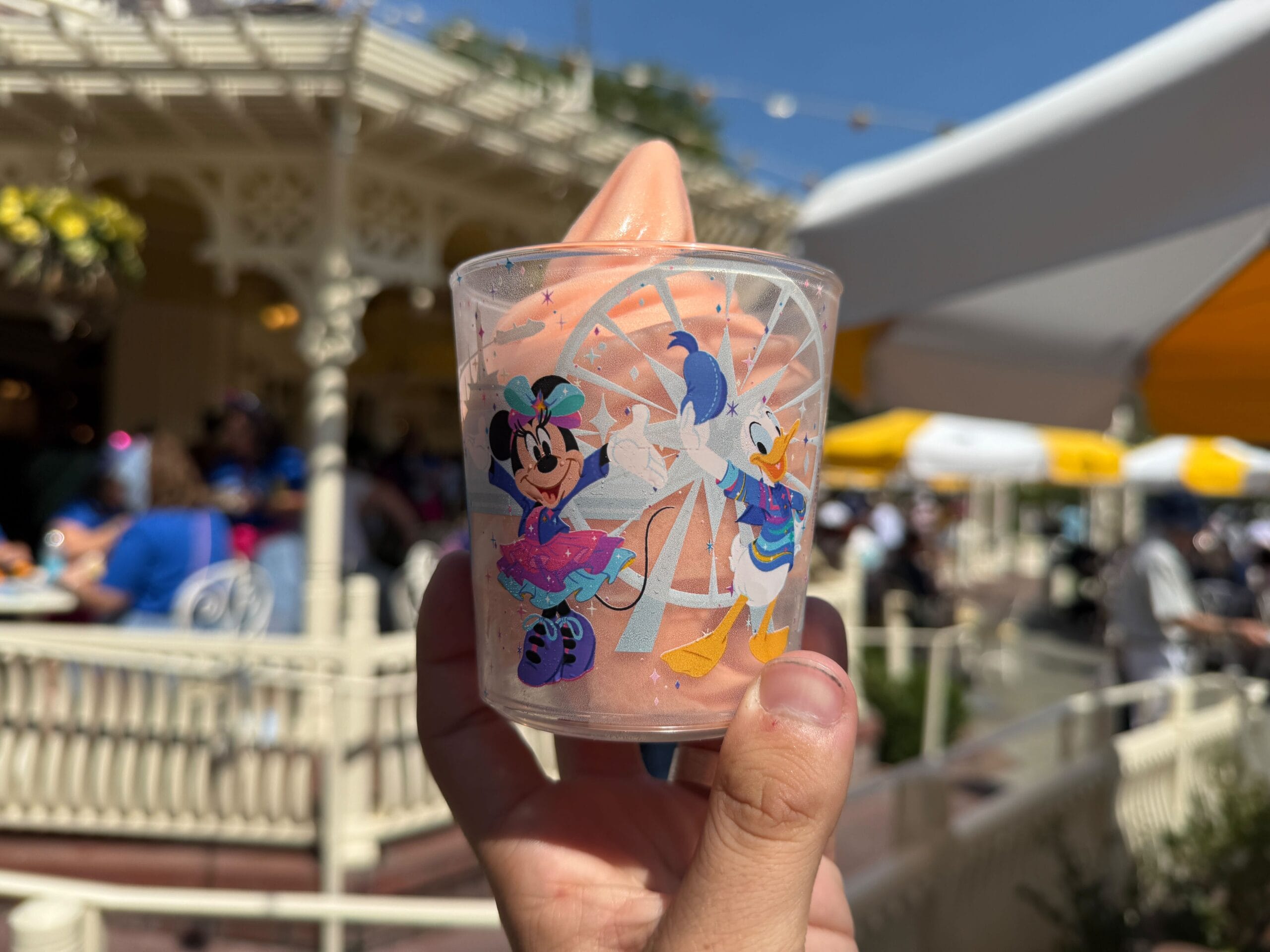 A hand holds an orange soft serve in a cup decorated with Minnie Mouse and Donald Duck at an outdoor venue.