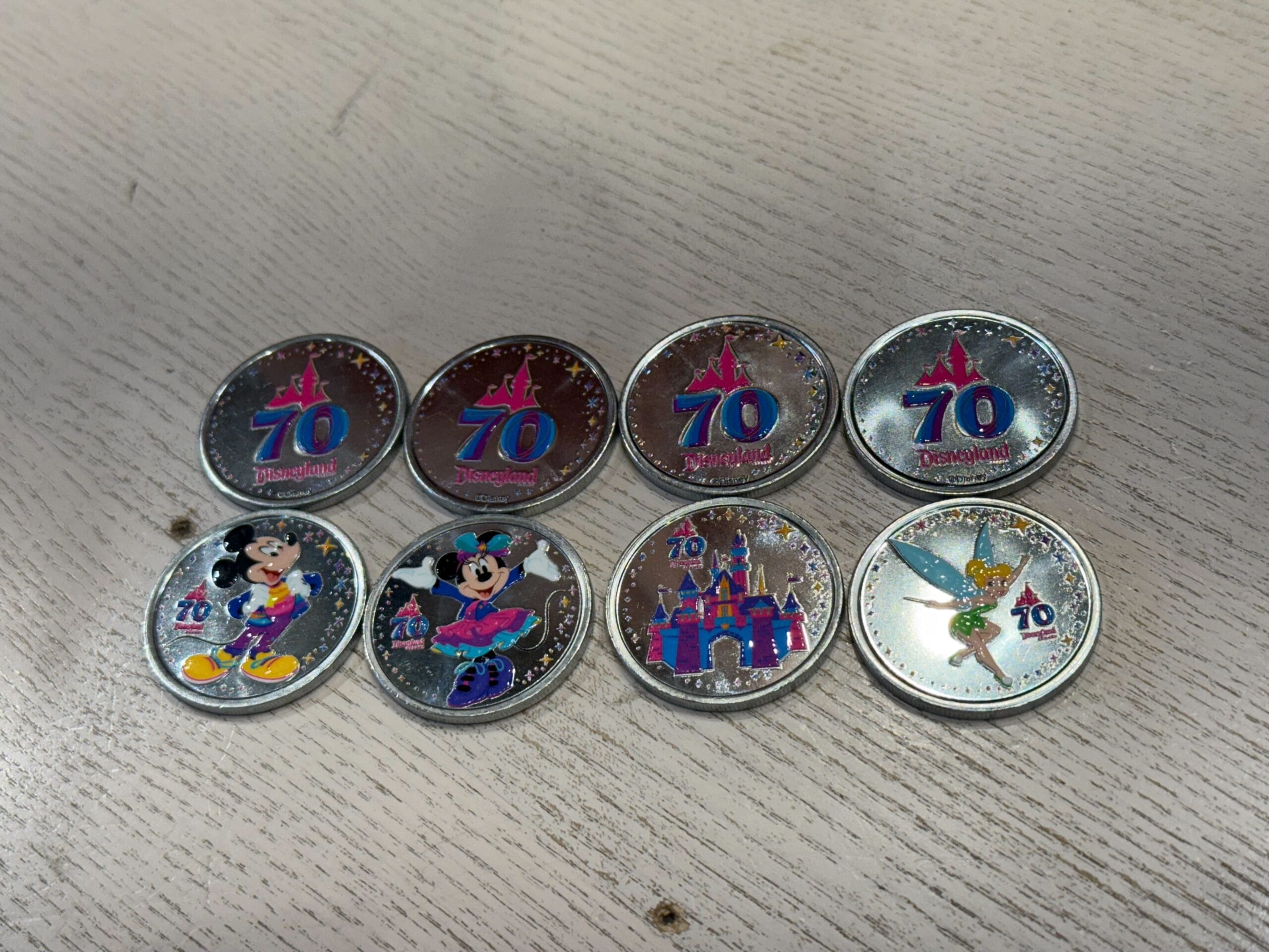 Eight Disneyland 70th anniversary collectible coins featuring characters and the castle on a light wood surface.