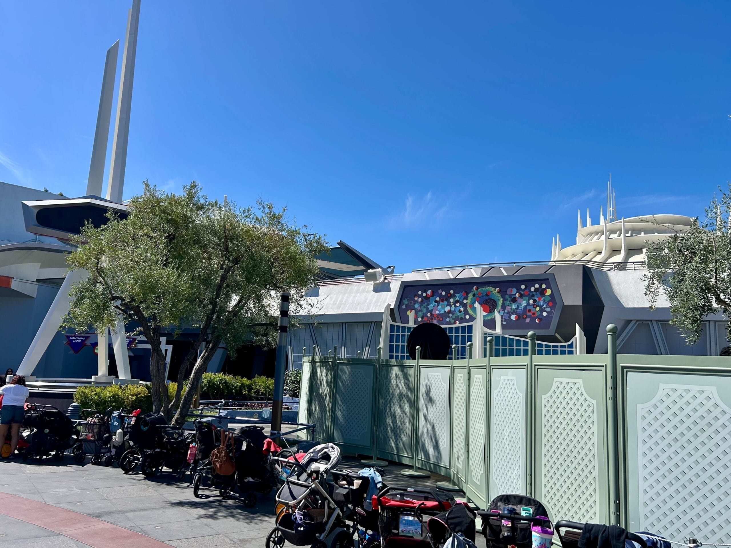 Green construction walls in front of a building at Disneyland Park, with strollers parked nearby under a clear blue sky.