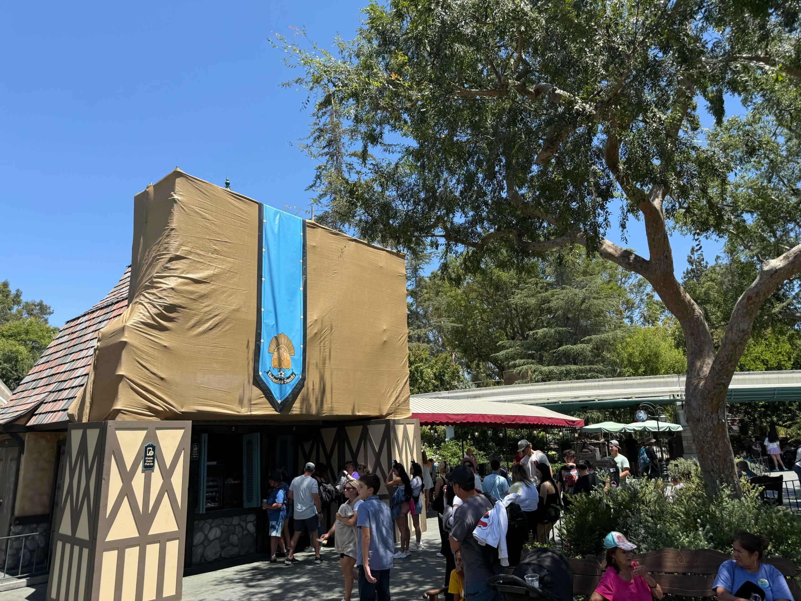 A small new construction covered in brown tarp with a blue banner, next to a line of people under a sunny sky.