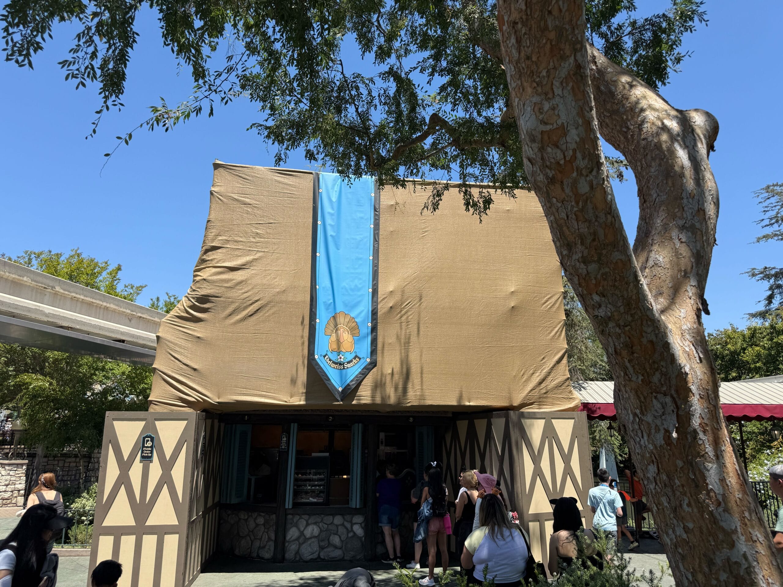 A new construction building covered in tan tarps with a blue banner; people stand outside under a sunny sky and trees.