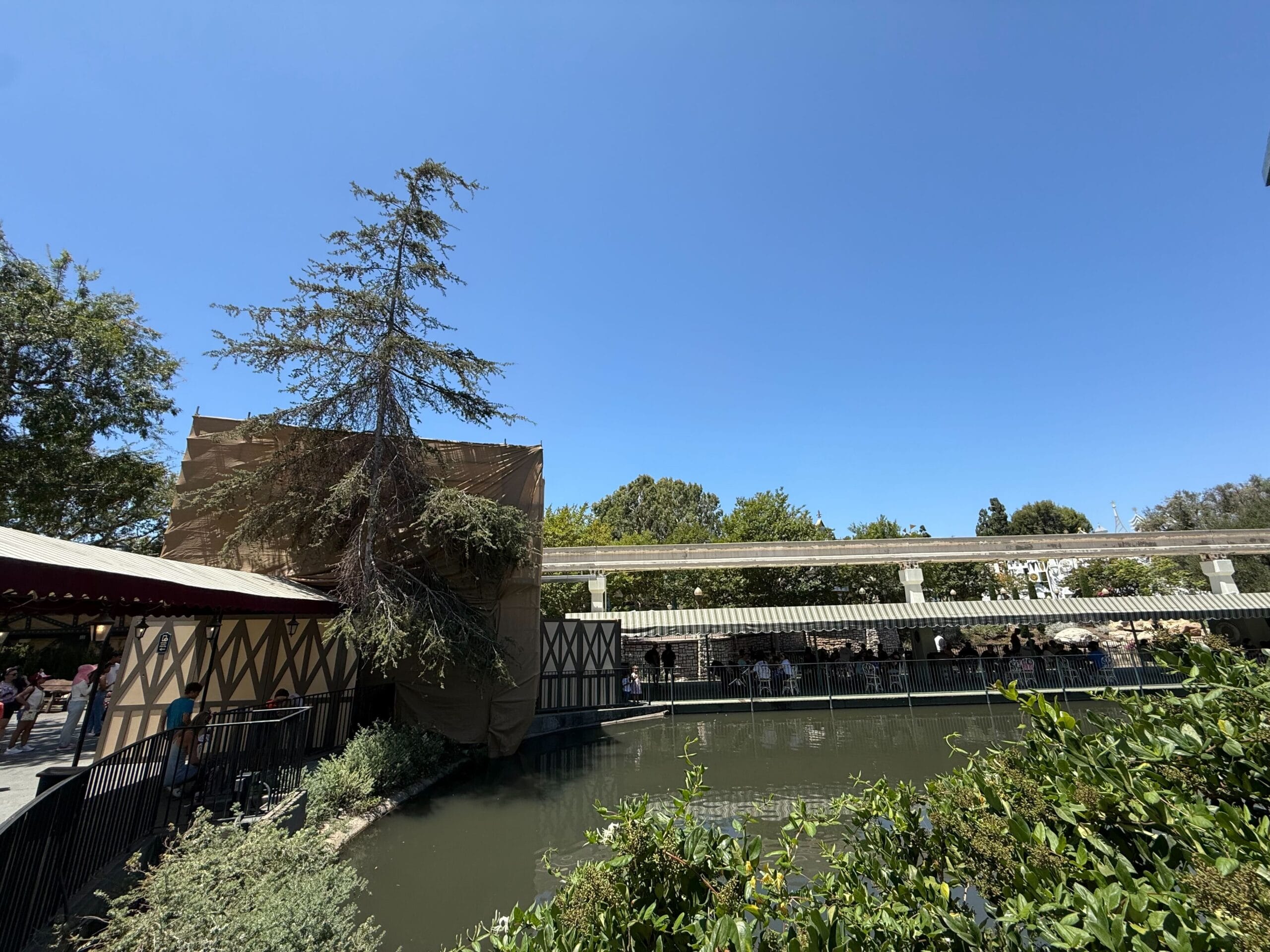 A pond surrounded by greenery, with a new construction building and people sitting under umbrellas on a sunny day.