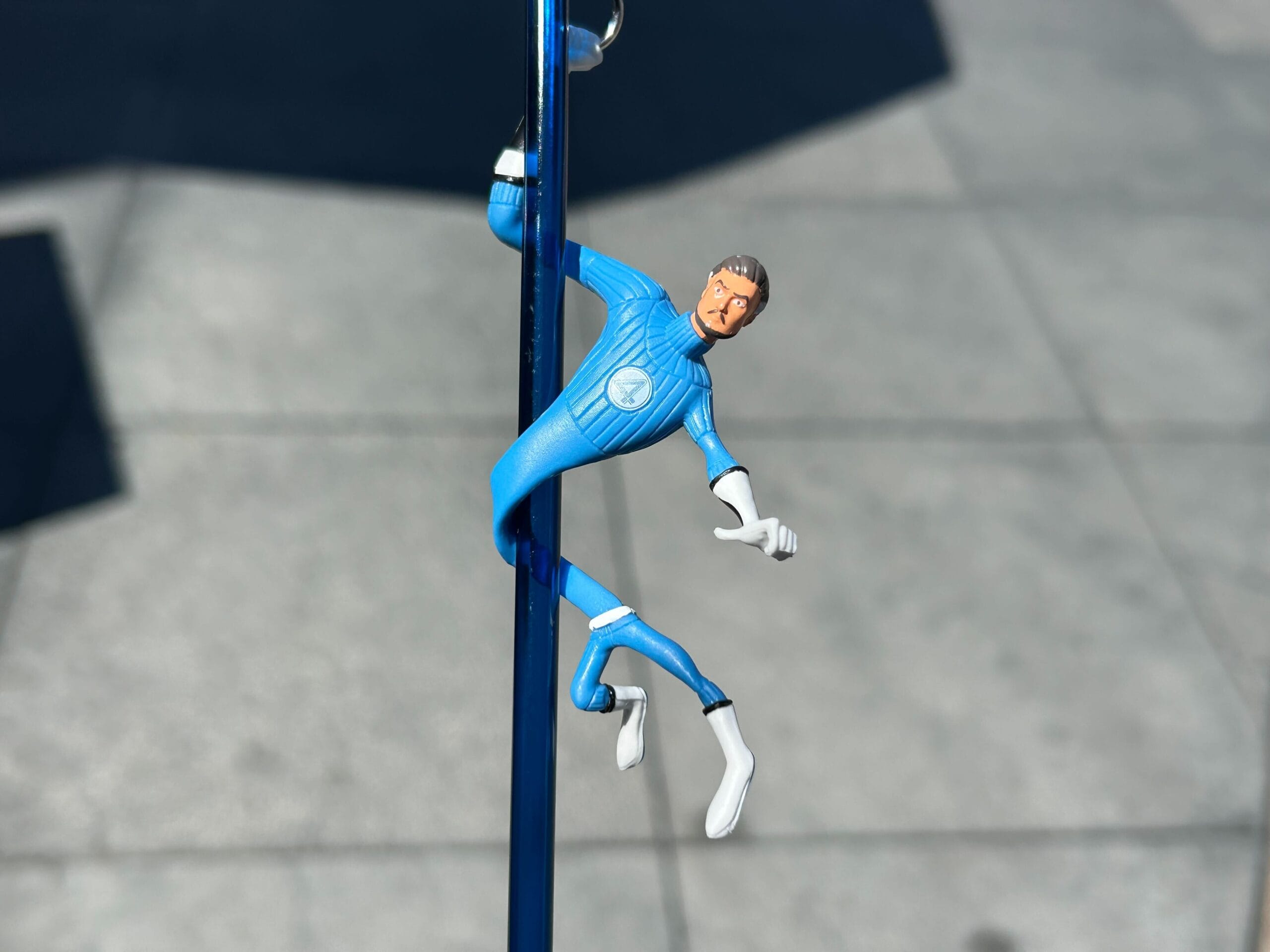 A stretchy action figure in a blue suit is hanging onto a vertical blue pole outdoors.