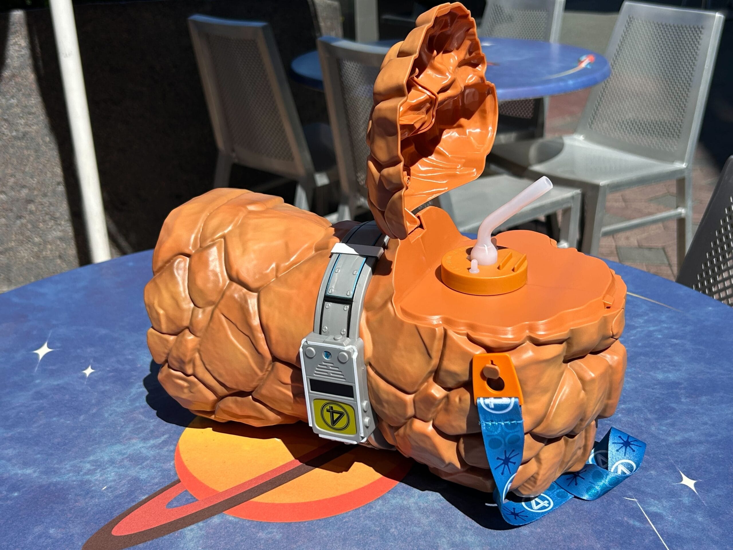 Plastic cup shaped like The Thing’s rocky arm, with a straw and lanyard, on an outdoor table with a space design.