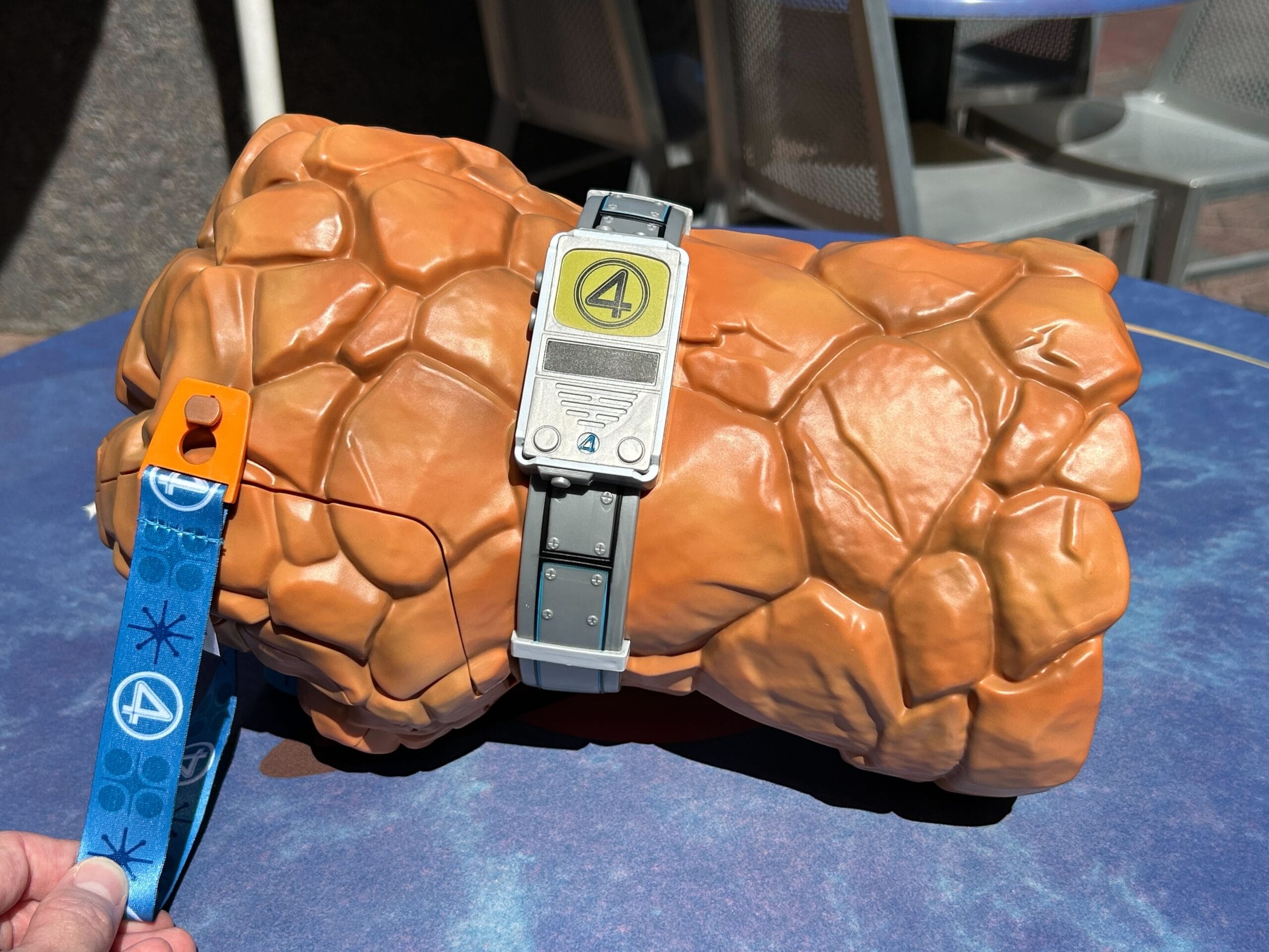A popcorn bucket shaped like The Thing's rocky hand with a Fantastic Four logo wristband, on a blue table.