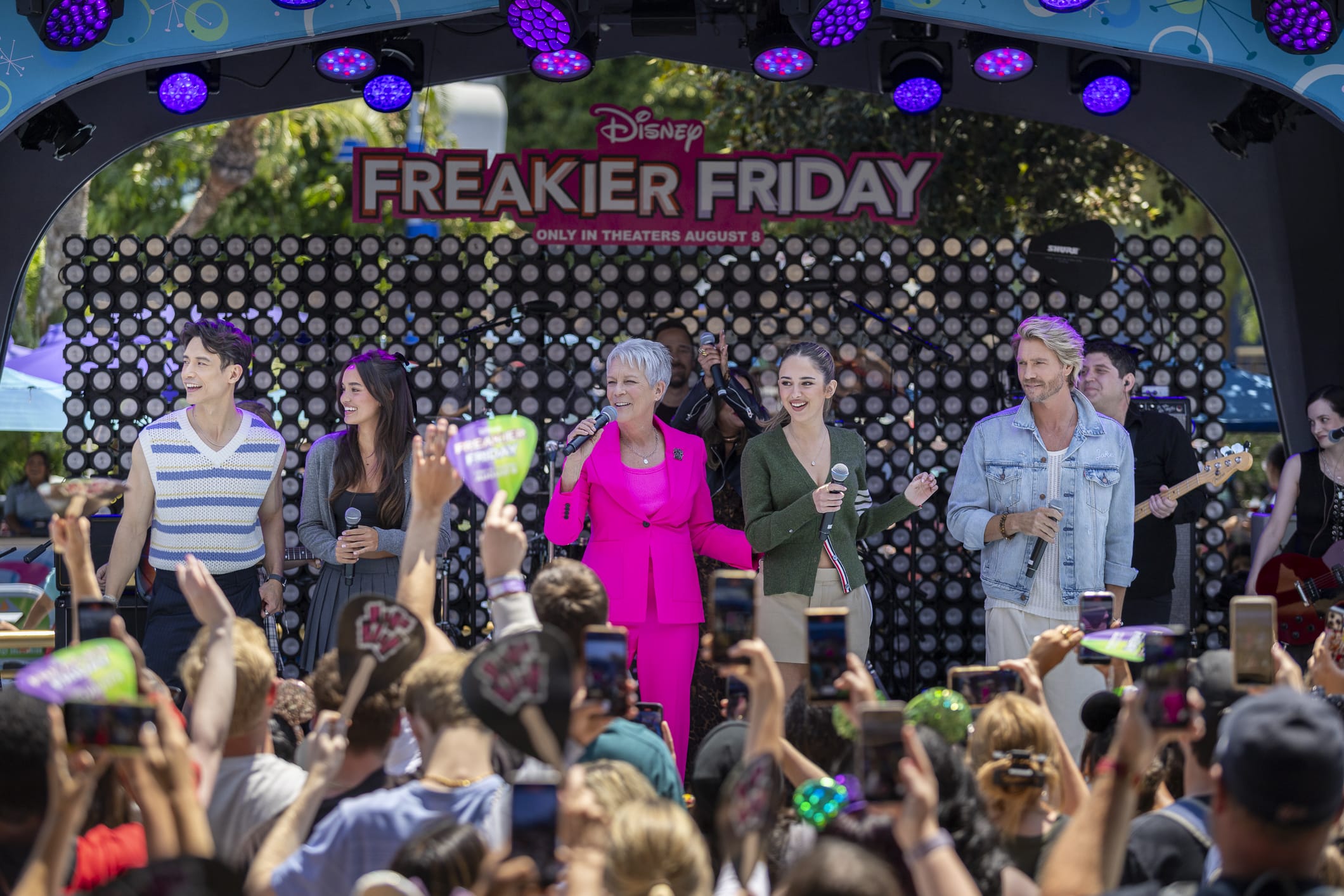 Freakier Friday cast on stage at a Disneyland event, delighting a crowd with phones and heart signs.