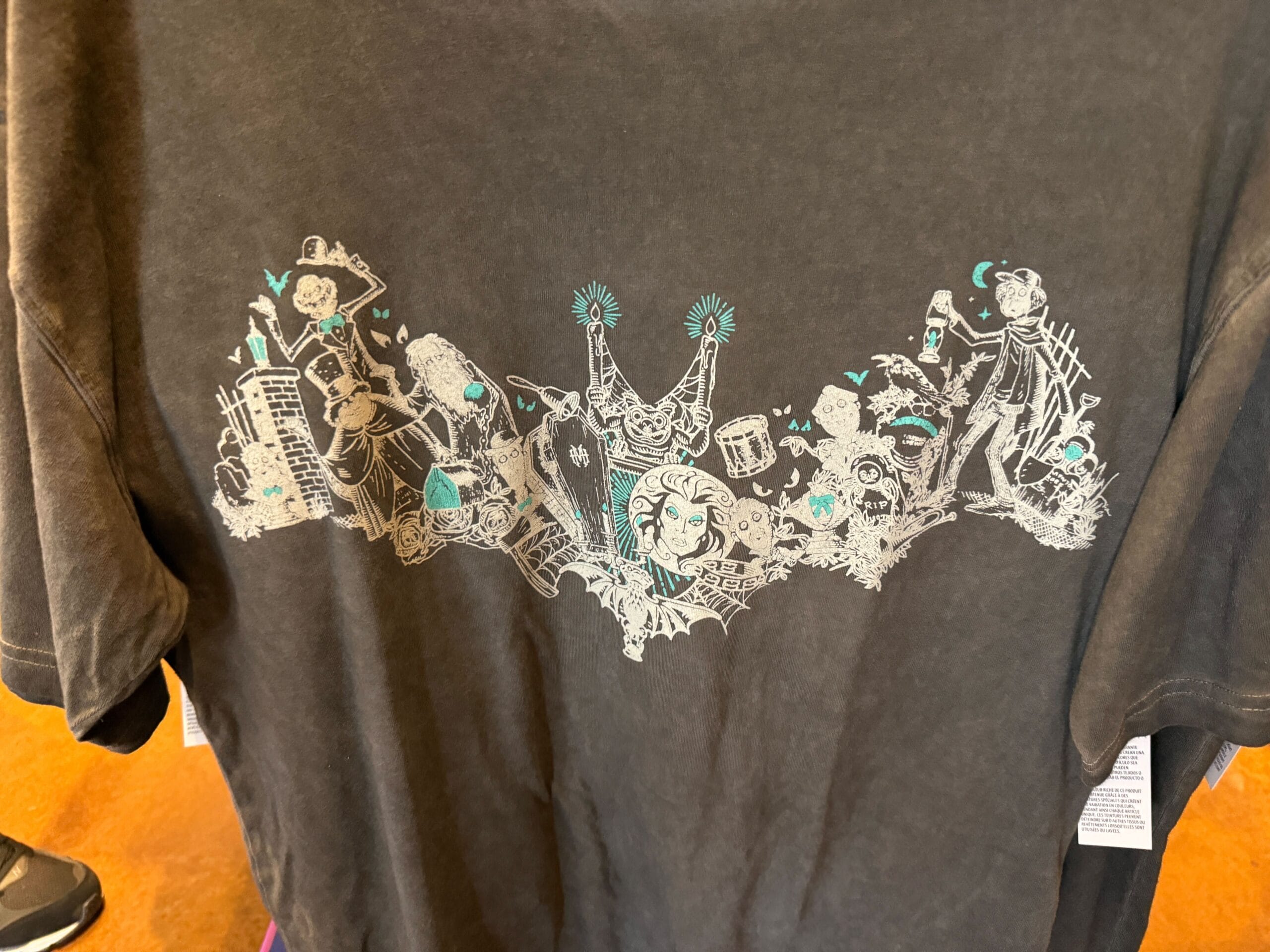 Black t-shirt with a white and teal graphic of musicians, dancers, and haunted mansion scenes in a lively, cartoonish style.