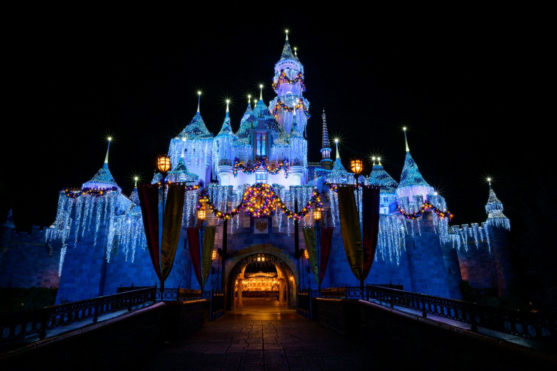 Disneyland Resort castle at night, glowing with festive lights and 2025 Holidays garlands in the dark.