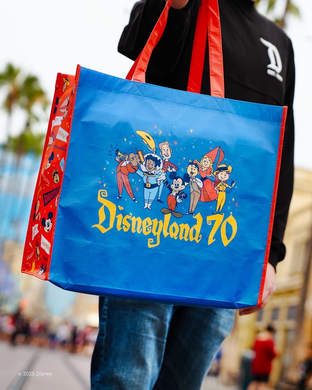 Person holding a blue Disneyland 70th anniversary tote bag with cartoon characters and red handles.