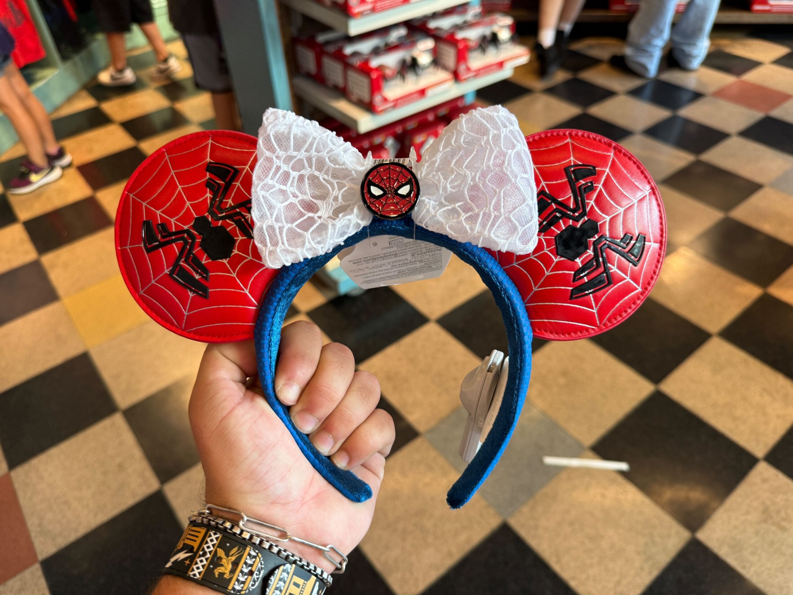 A hand holds Spider-Man themed Minnie Mouse ear headband with a white lace bow in a store.