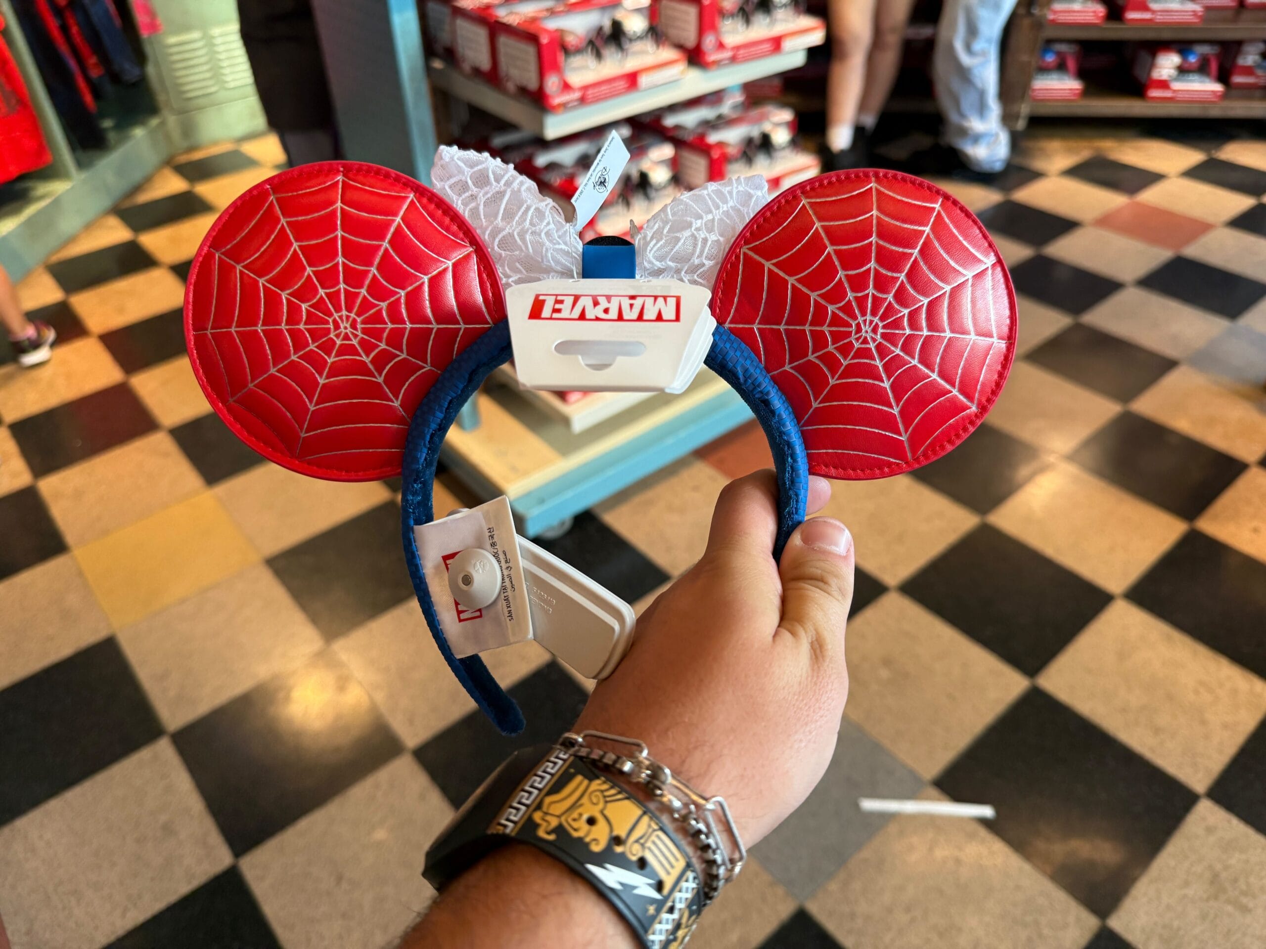 A hand holds Marvel Spider-Man themed Minnie Mouse ears headband in a store with checkered floor tiles.