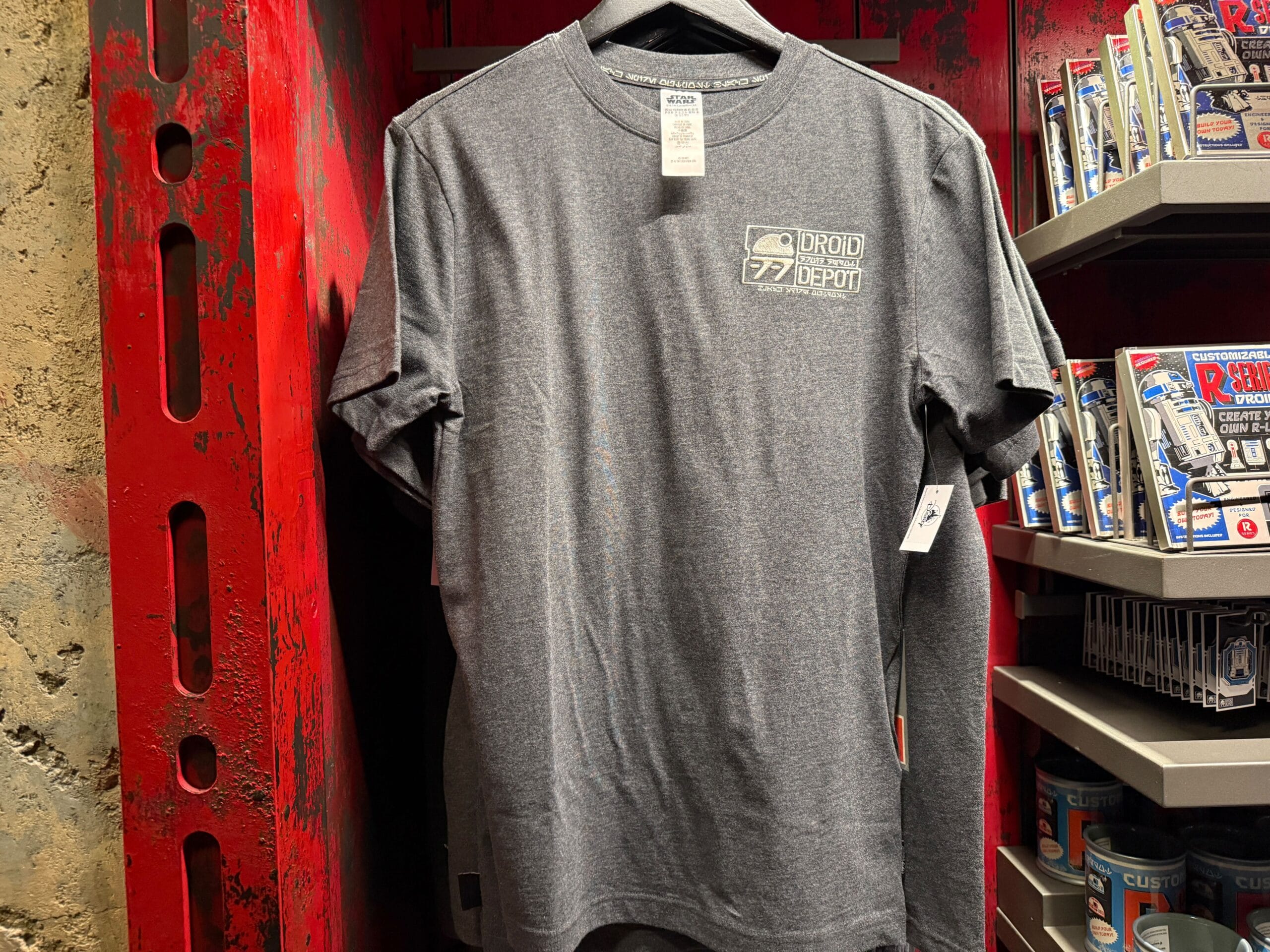 Gray "Droid Depot" t-shirt hangs on a rack in a store with shelves of merchandise in the background.