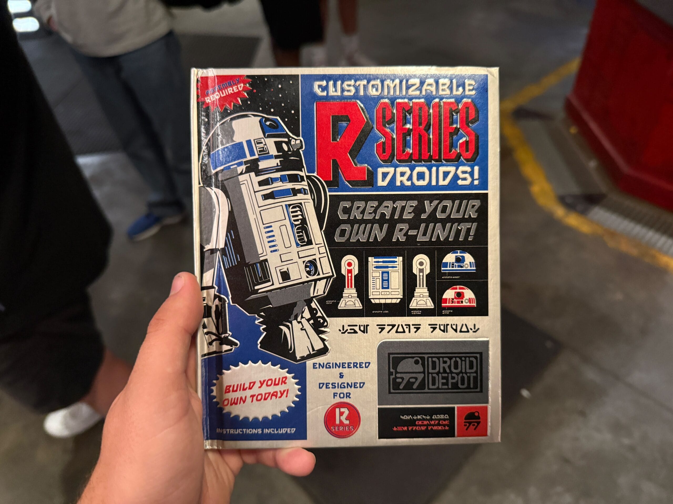A hand holds a "Customizable R-Series Droids" booklet featuring R2-D2 and droid-building graphics.