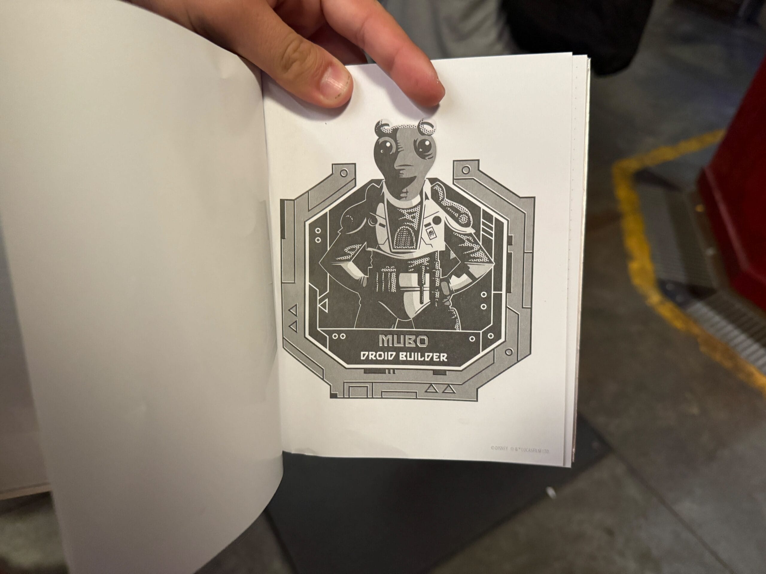 A hand holds a book open to a page showing a drawing labeled "Milbo Droid Builder" with a robot character.