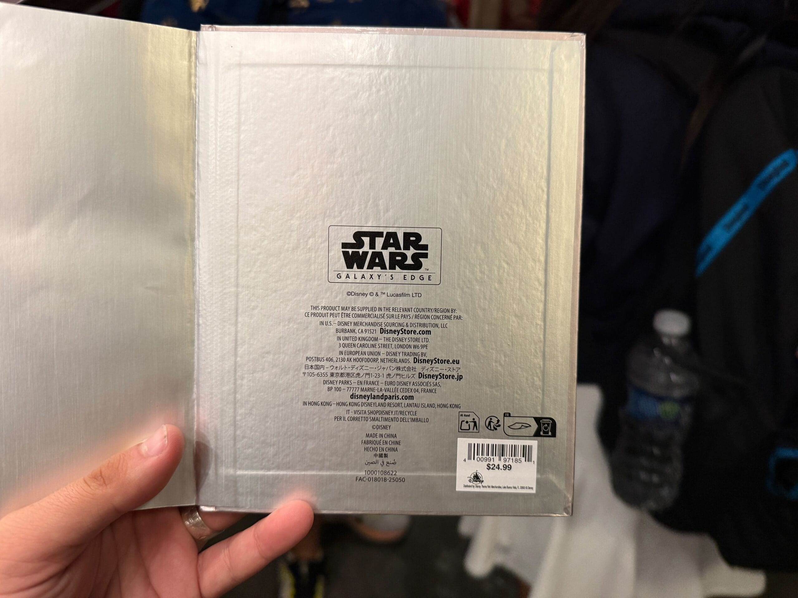 A hand holds a "Star Wars: Galaxy's Edge" item with a $29.99 price tag, showing text and logos on the back cover.