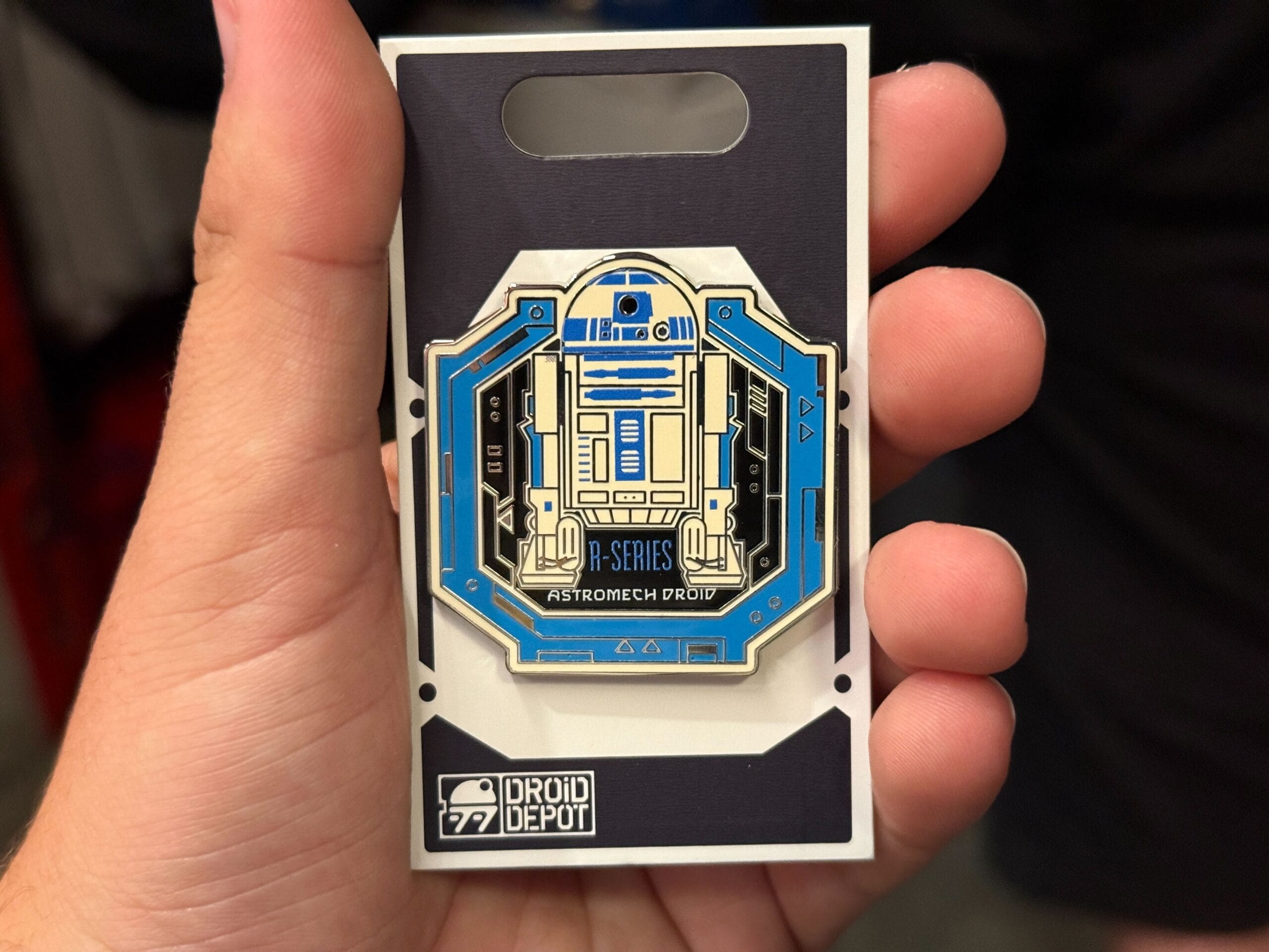 A hand holds a Droid Depot pin featuring the blue and white R2-series astromech droid, R2-D2.