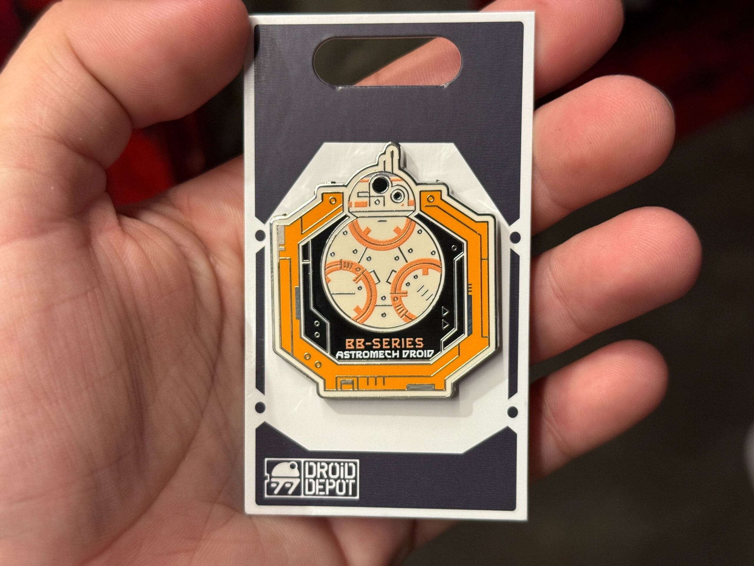 A hand holds a BB-Series Astromech Droid pin from Droid Depot, featuring orange and white droid artwork.