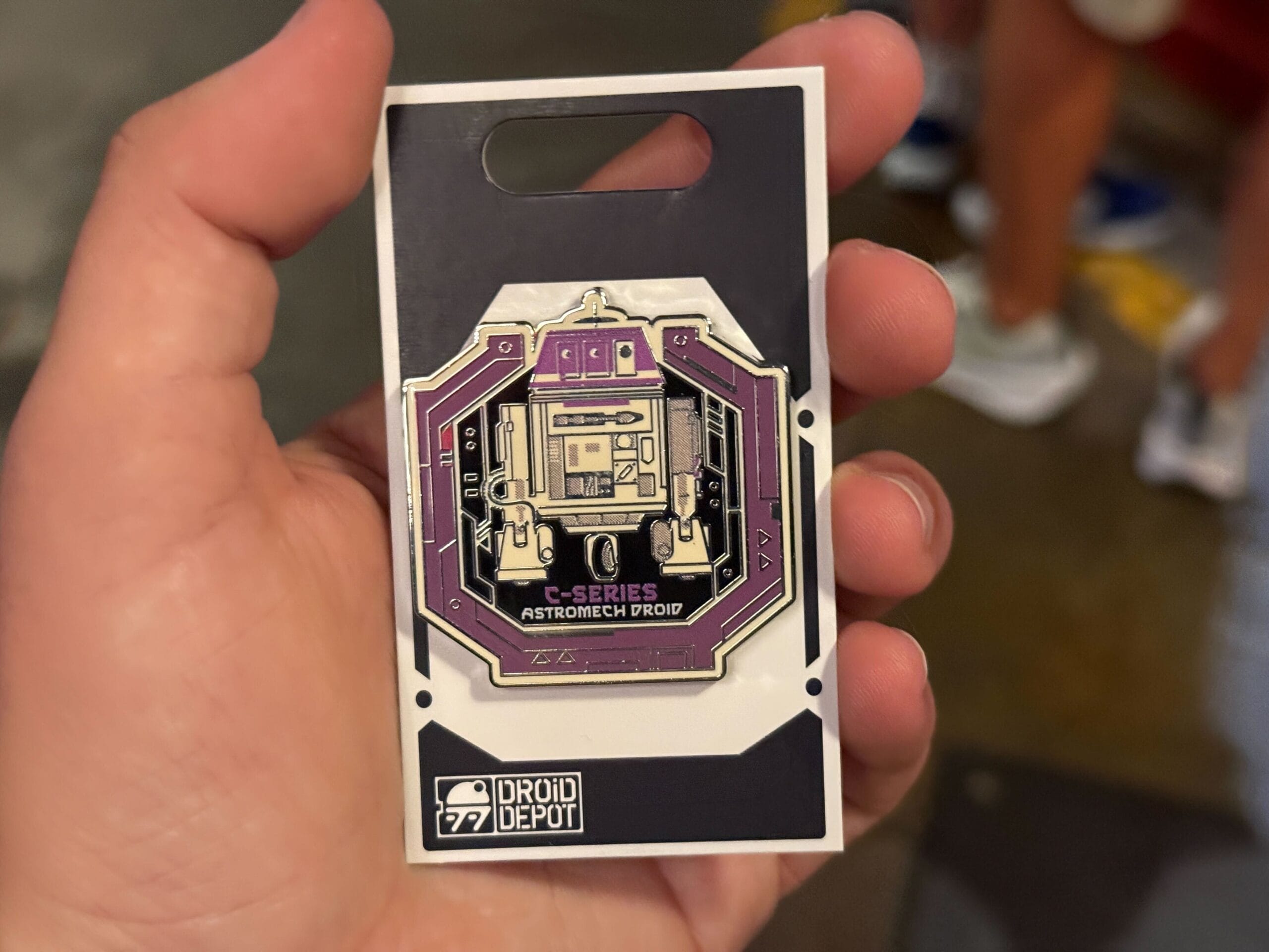 A hand holds a C-Series Astromech Droid pin from Droid Depot, still attached to its display backing.