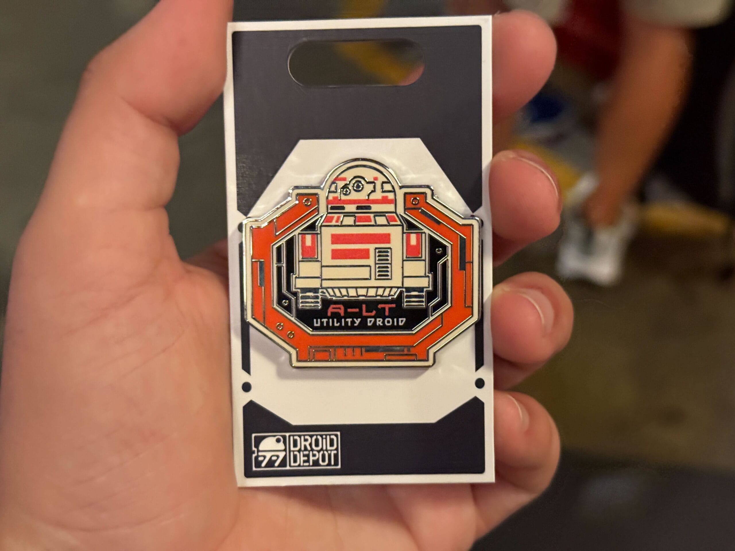 A hand holds a Droid Depot pin featuring a red and white A-LT utility droid on an orange background.