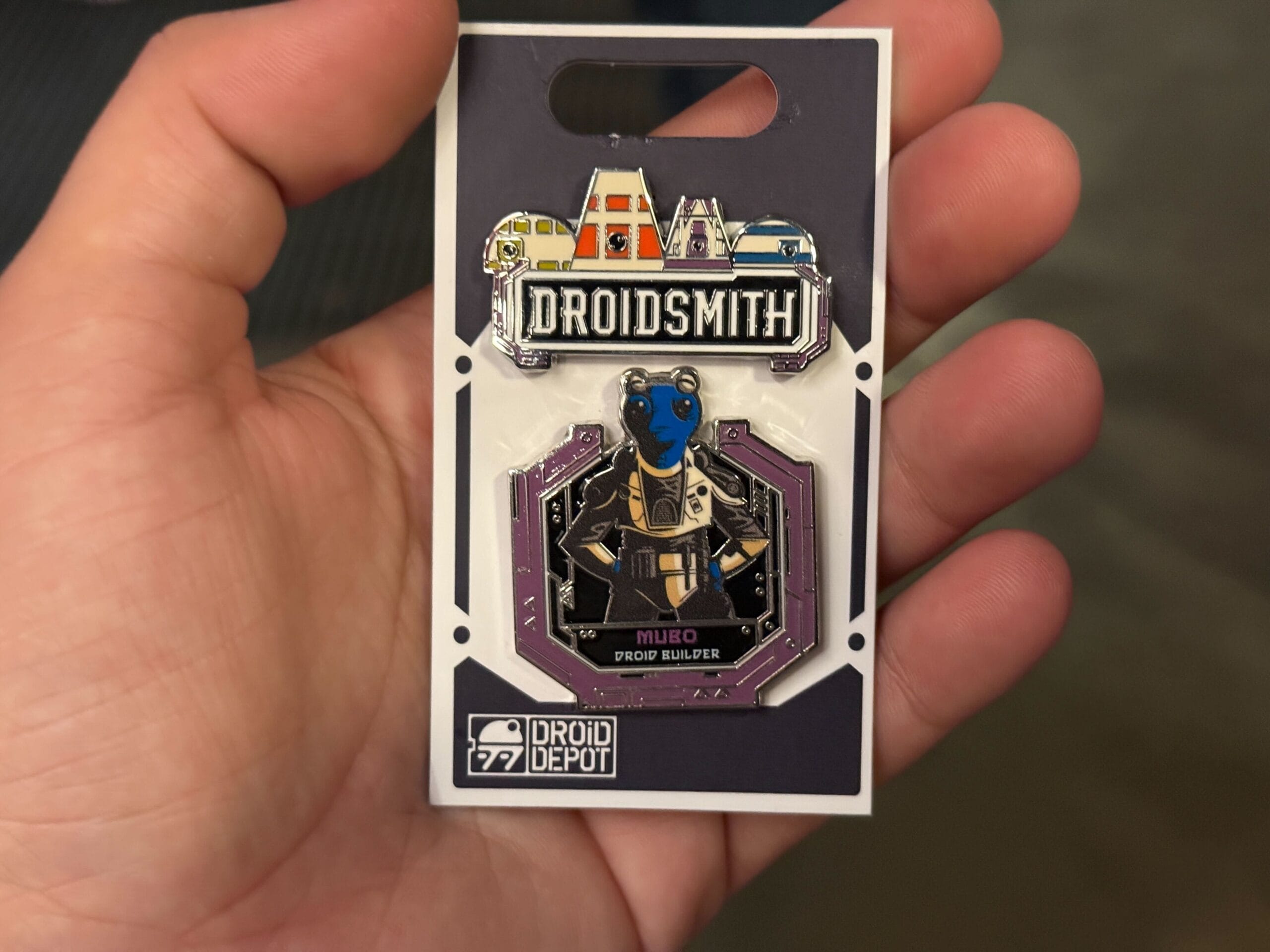 A hand holds a Droidsmith pin featuring a blue alien character named Milbo from Droid Depot.