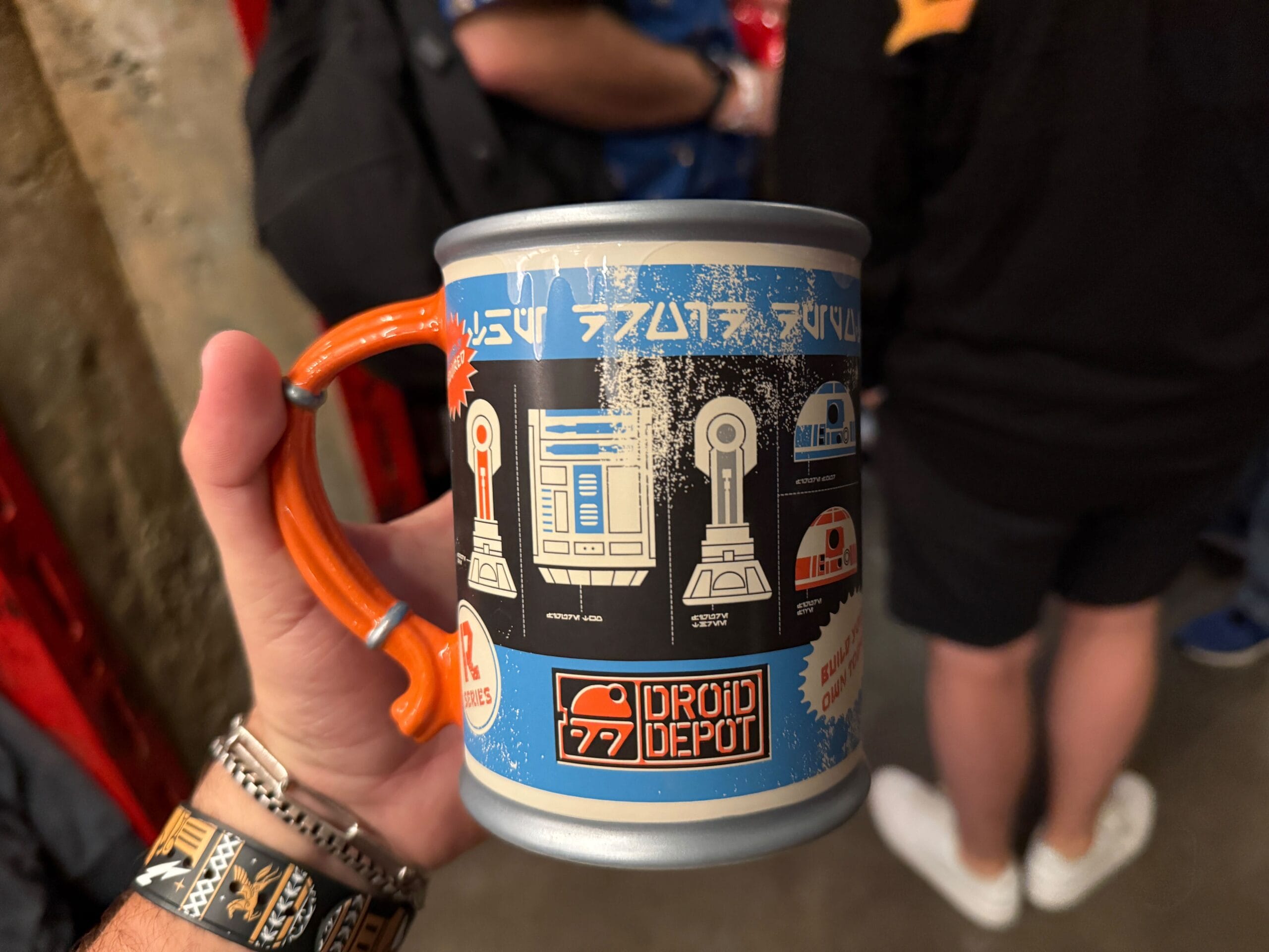 A hand holds a Droid Depot mug featuring Star Wars droid designs and blue, orange, and black graphics.
