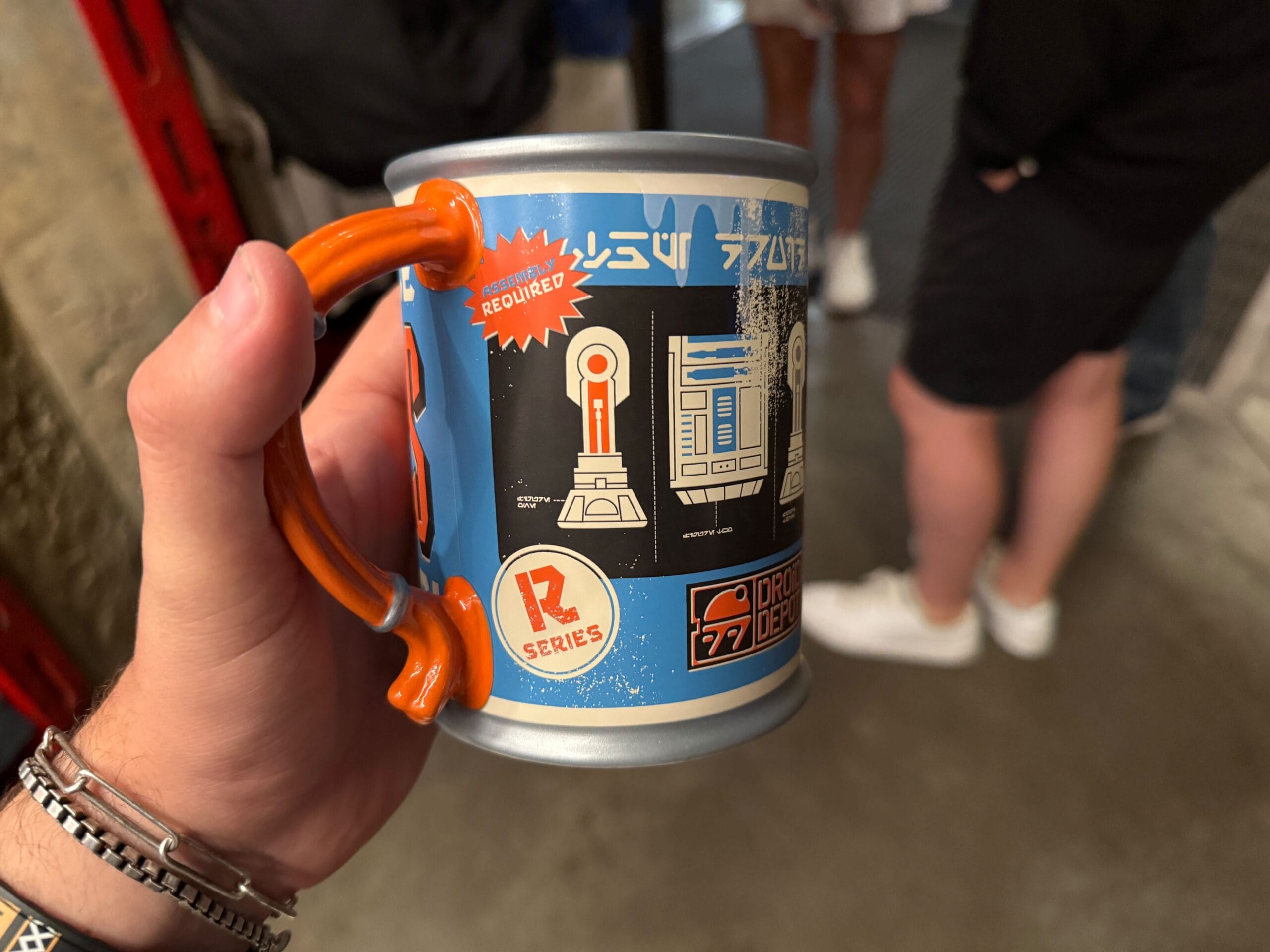 A hand holds a blue Star Wars mug with droid graphics and text; people stand blurred in the background.