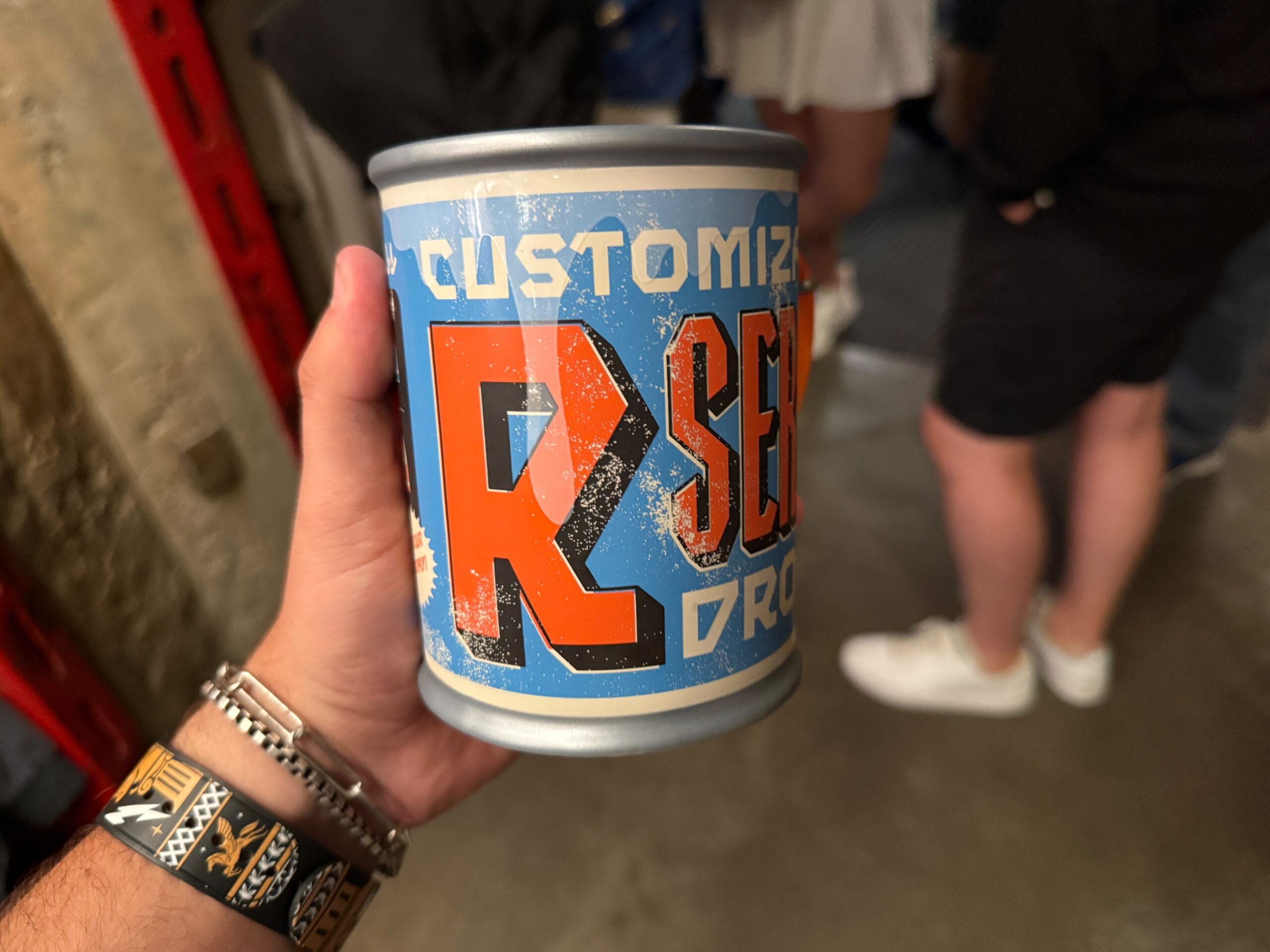 A person holds a can with colorful, bold text that reads "CUSTOMIZE" in a crowded indoor setting.