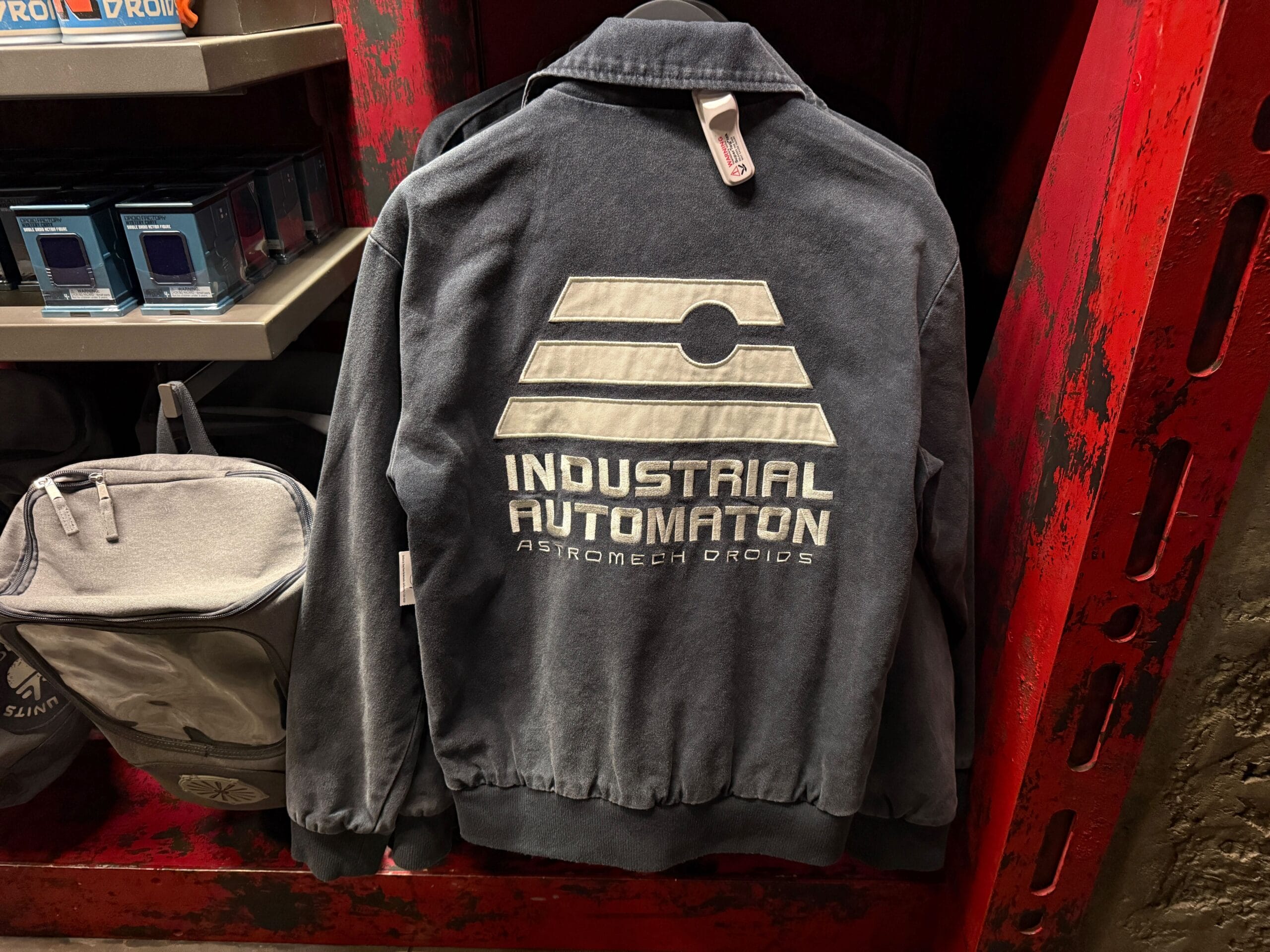 A gray jacket with "Industrial Automaton Astromech Droids" and a logo printed on the back, hanging on a rack.