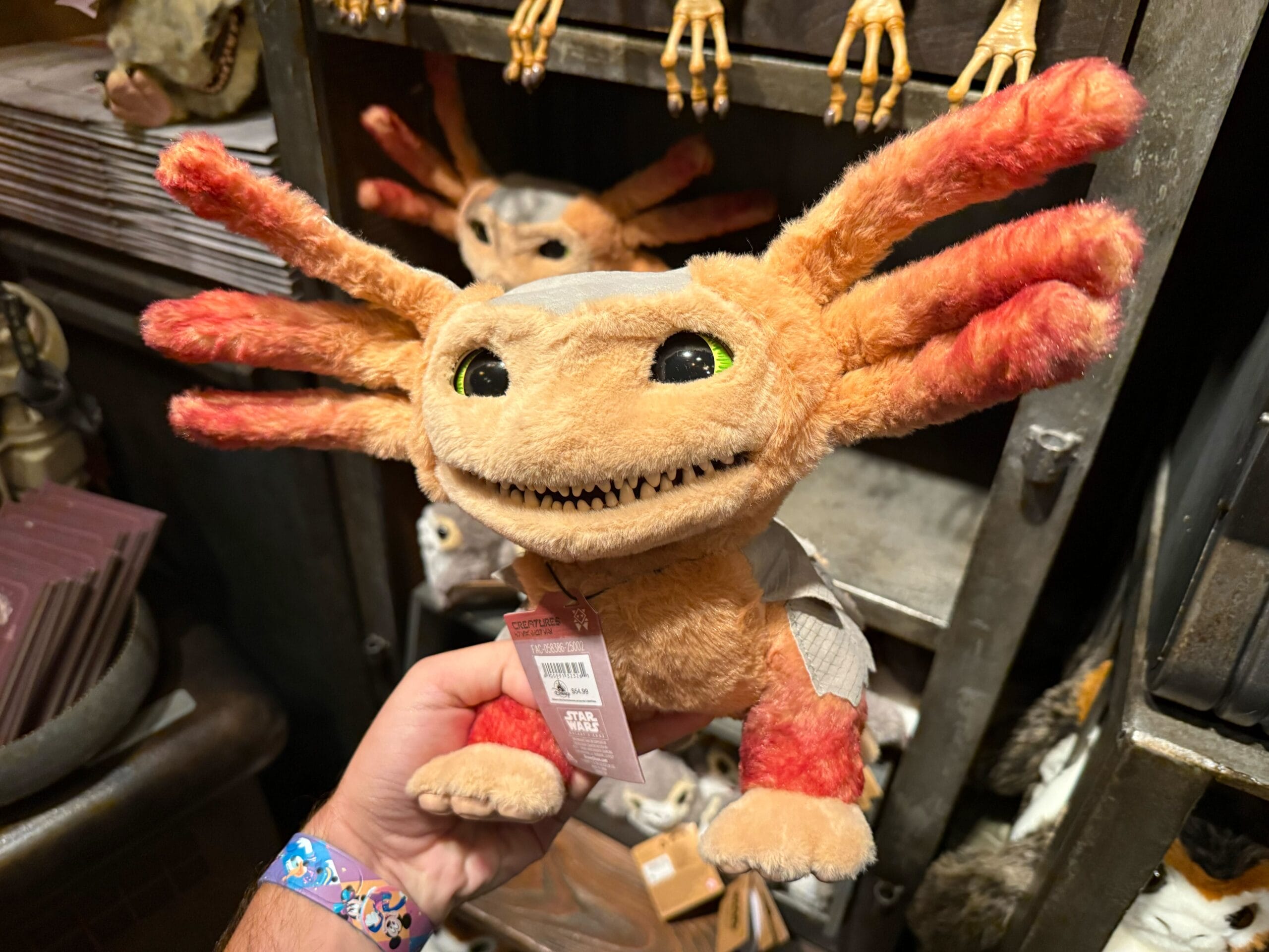 A hand holds a plush toy creature with orange-red limbs, yellow eyes, and a wide, toothy smile in a shop.