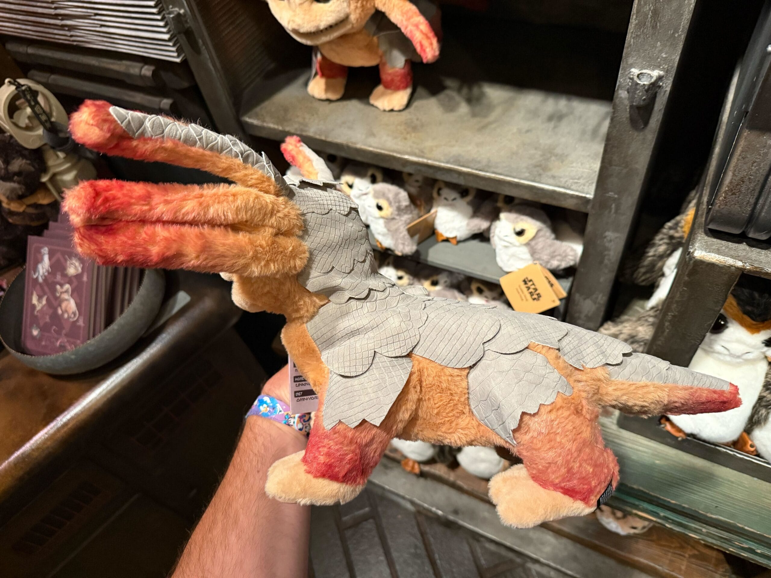 A hand holds a plush toy creature with orange fur and gray scale-like armor, displayed in a store.
