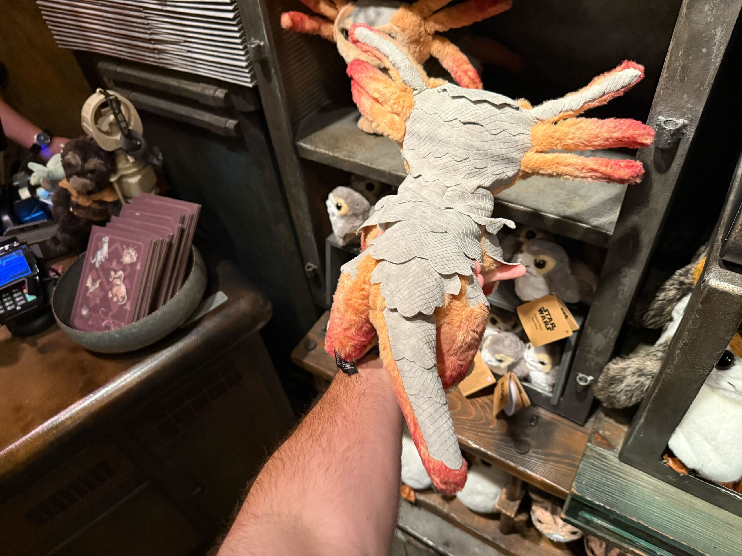 A plush toy resembling a phoenix is perched on a person's arm inside a store display with other stuffed animals.