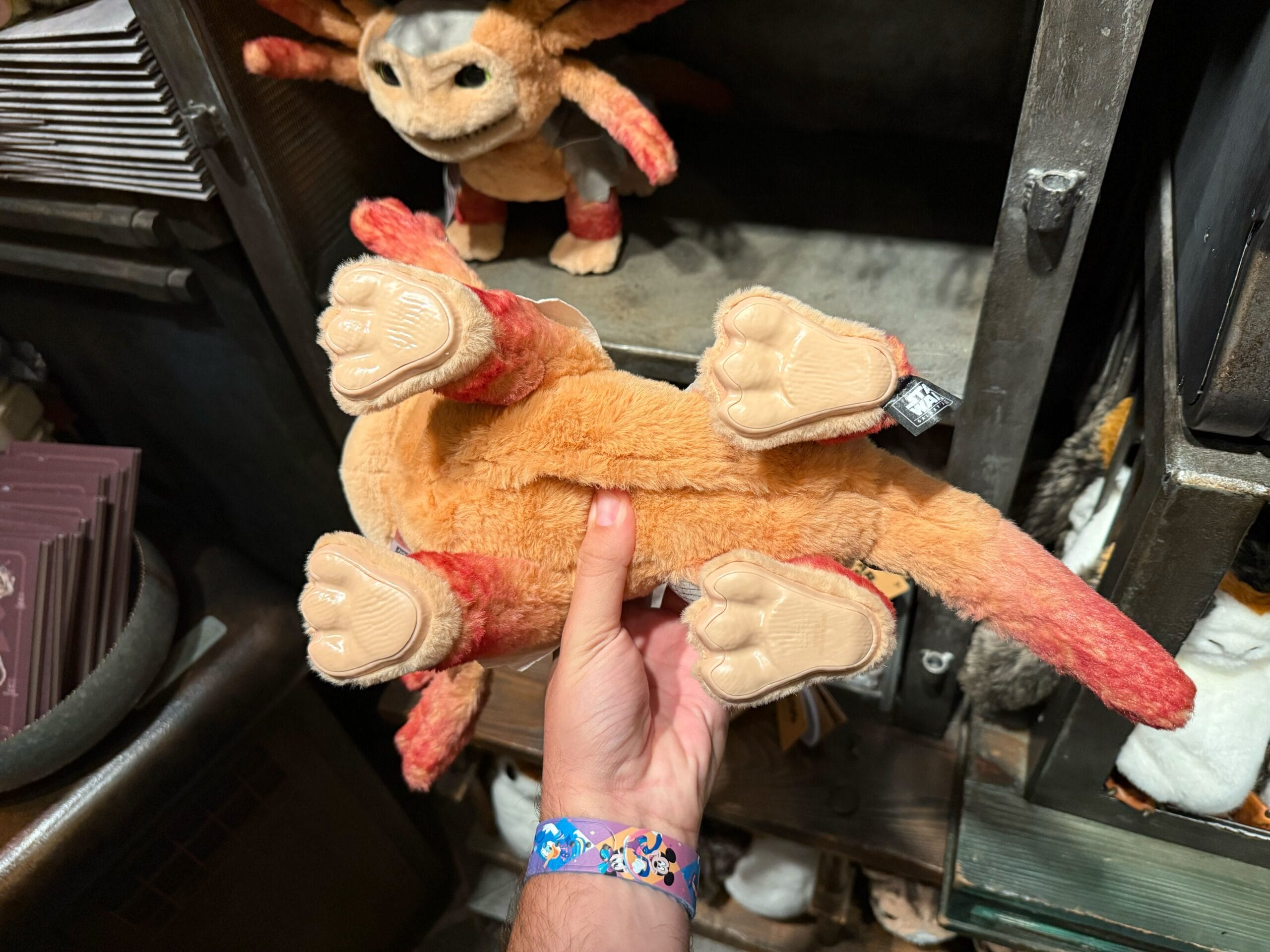 A hand holds a plush toy creature upside down, showing its four beige paws and orange fur.